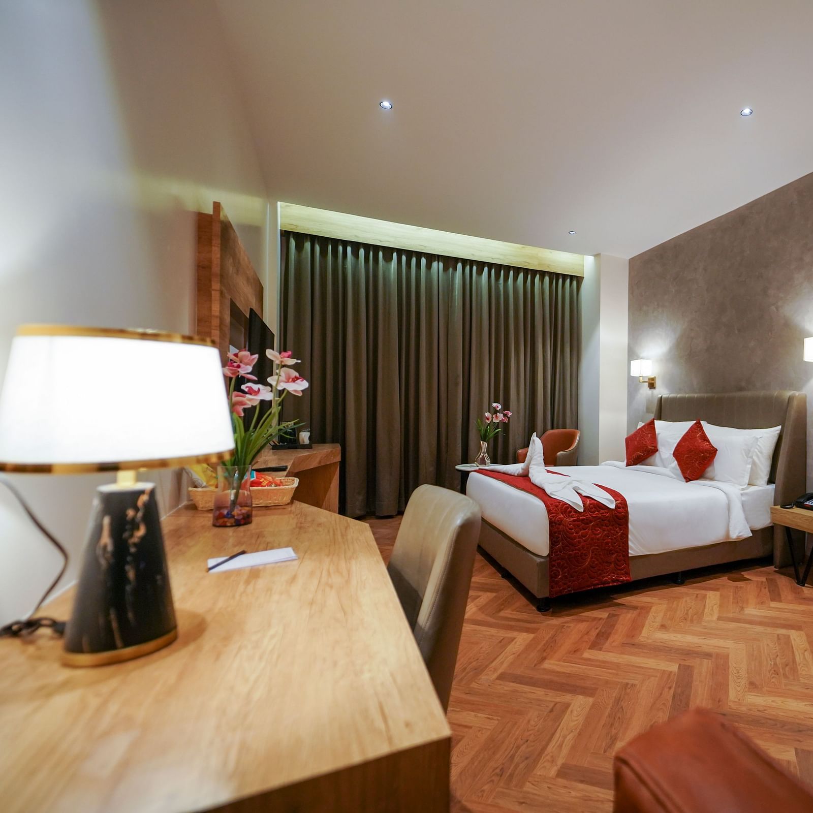 A well-lit hotel room includes a bed with pillows, a wooden desk with a lamp, a chair, curtains, and patterned flooring - Hotel Park Inn, Nellore.