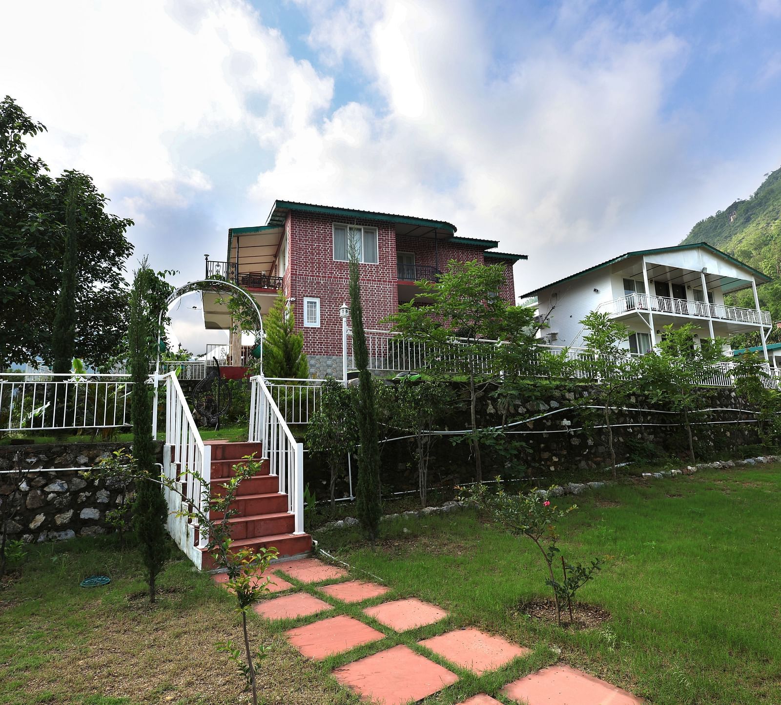 Resort buildings with red brick and white exteriors, set amidst green lawns, trees, and a stone pathway with white railings under a partly cloudy sky - Neer Ganga Resorts