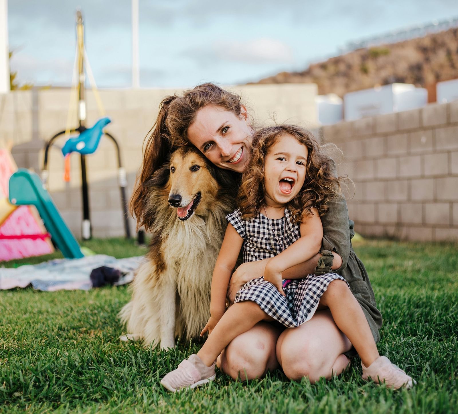 A smiling woman and a young girl with curly hair hug a fluffy dog on a green lawn with toys in the background.