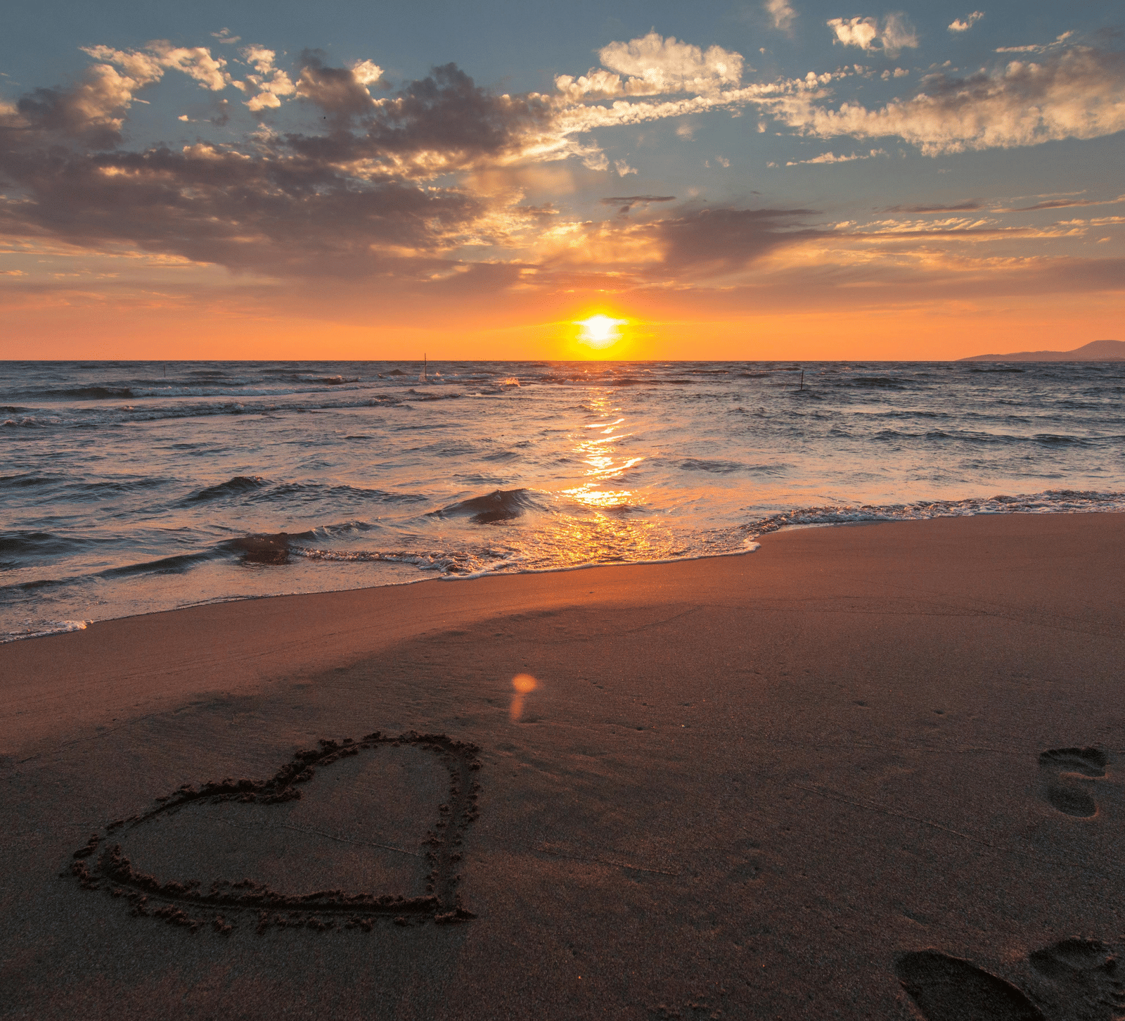Sunset view over a beach, with waves gently rolling onto the sand where a heart shape has been drawn in the foreground.