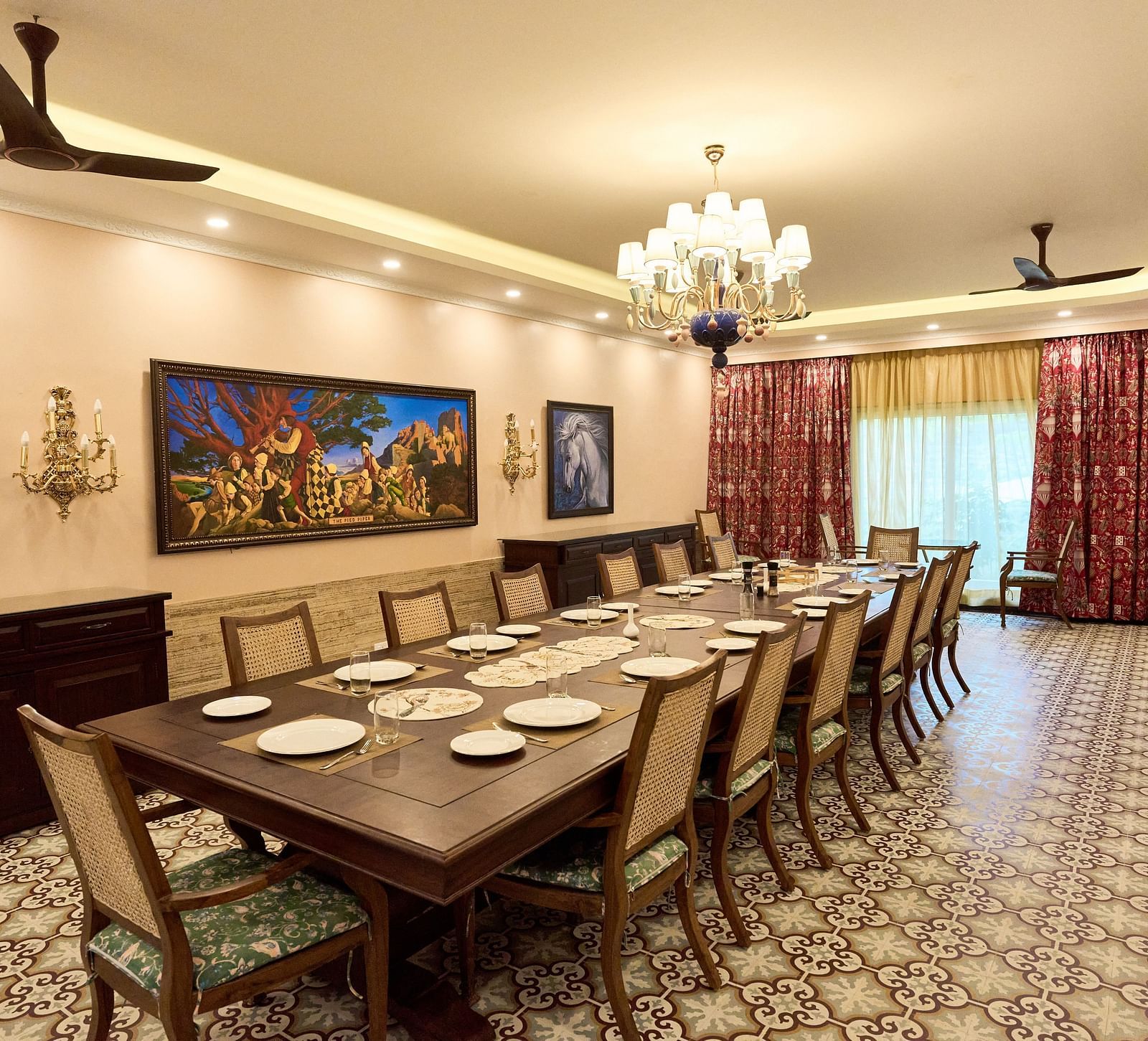 Diagonal view of a large dining table set for a culinary experience, with wall-mounted paintings, a chandelier, and large windows at Mayfair Manor, Alibag