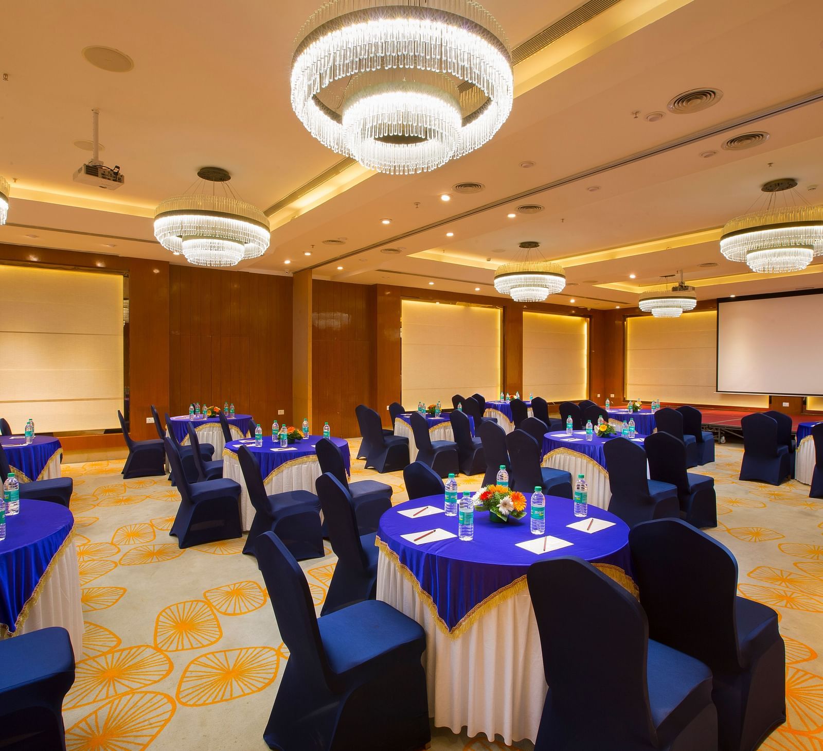 Another view of a large conference hall with circular tables, blue chairs and a projector screen, ready for a seminar - Hotel Southend by TGI - Bommasandra, Bangalore