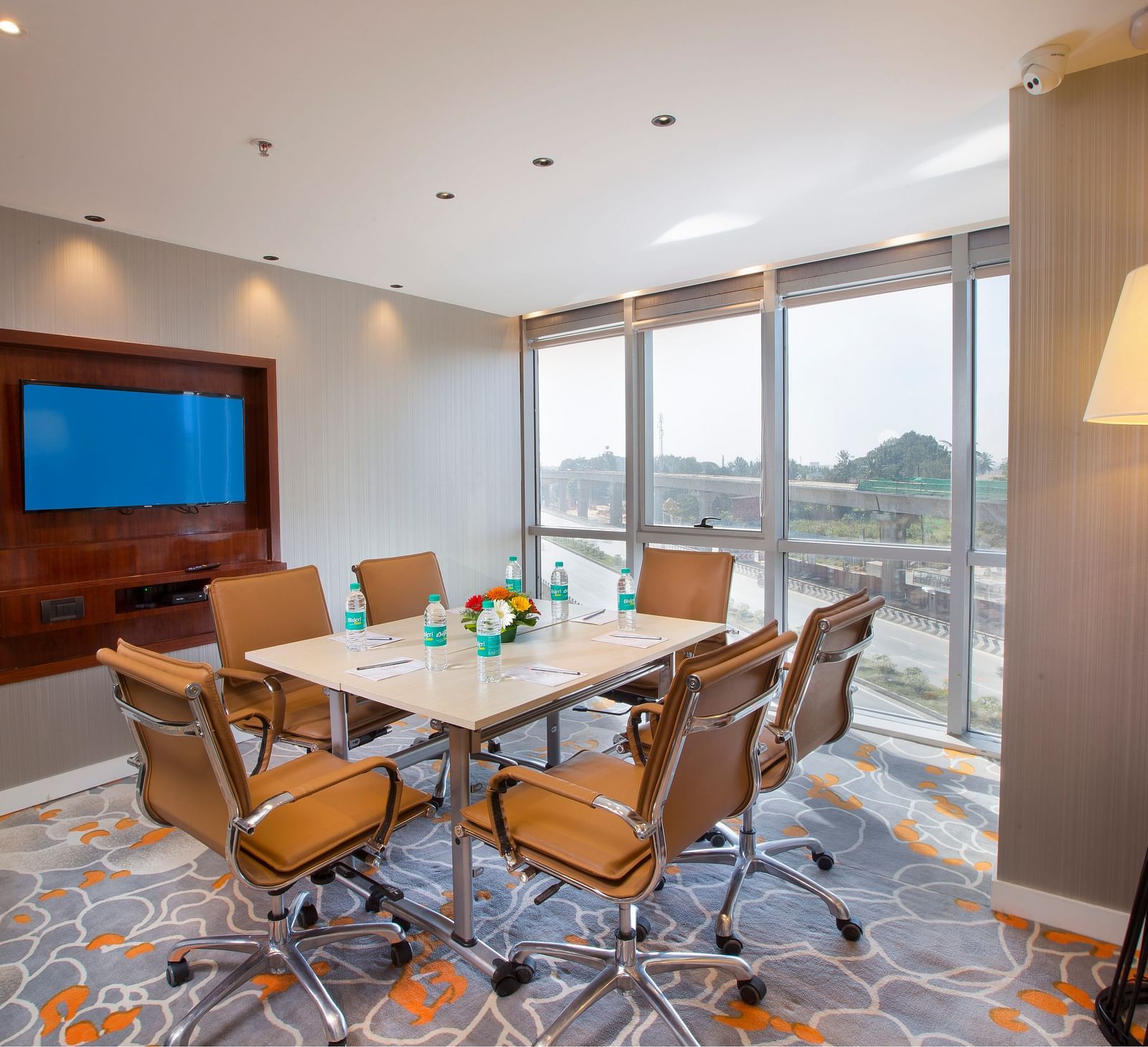 A small meeting room with a round table, tan leather chairs and a TV, with a large window and a floor lamp - Hotel Southend by TGI - Bommasandra, Bangalore