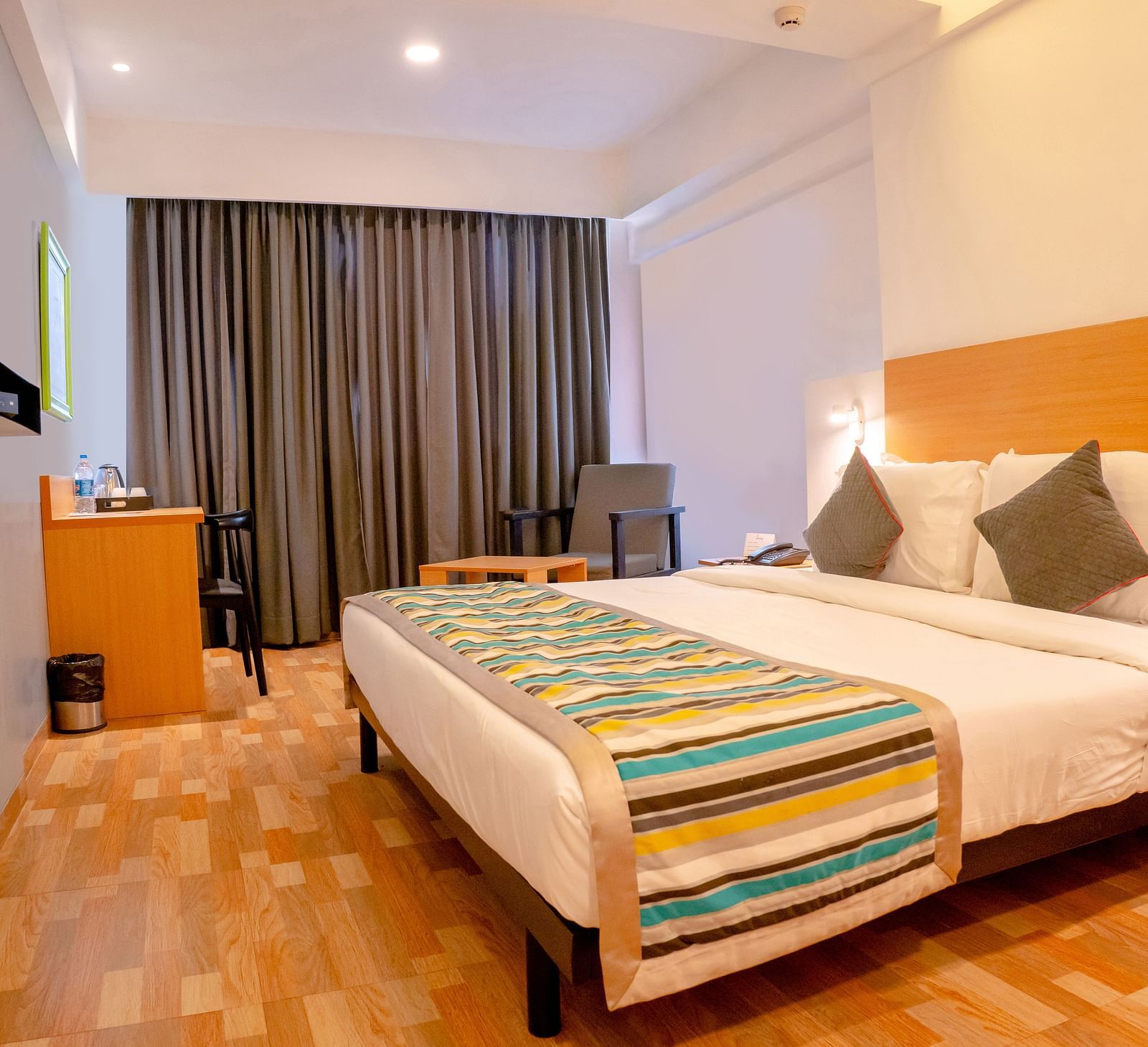 Another view of a cosy hotel room with a large bed, colourful throw and wooden furniture, including a desk with a TV above it - TGI Ameya Grand, Goa