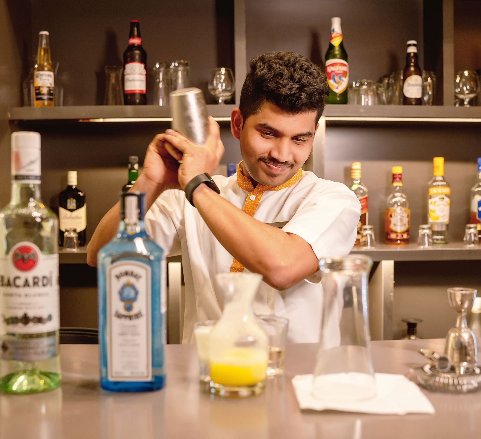 A bartender shaking a cocktail with bottles of alcohol in the background - TGI Ameya Grand, Goa