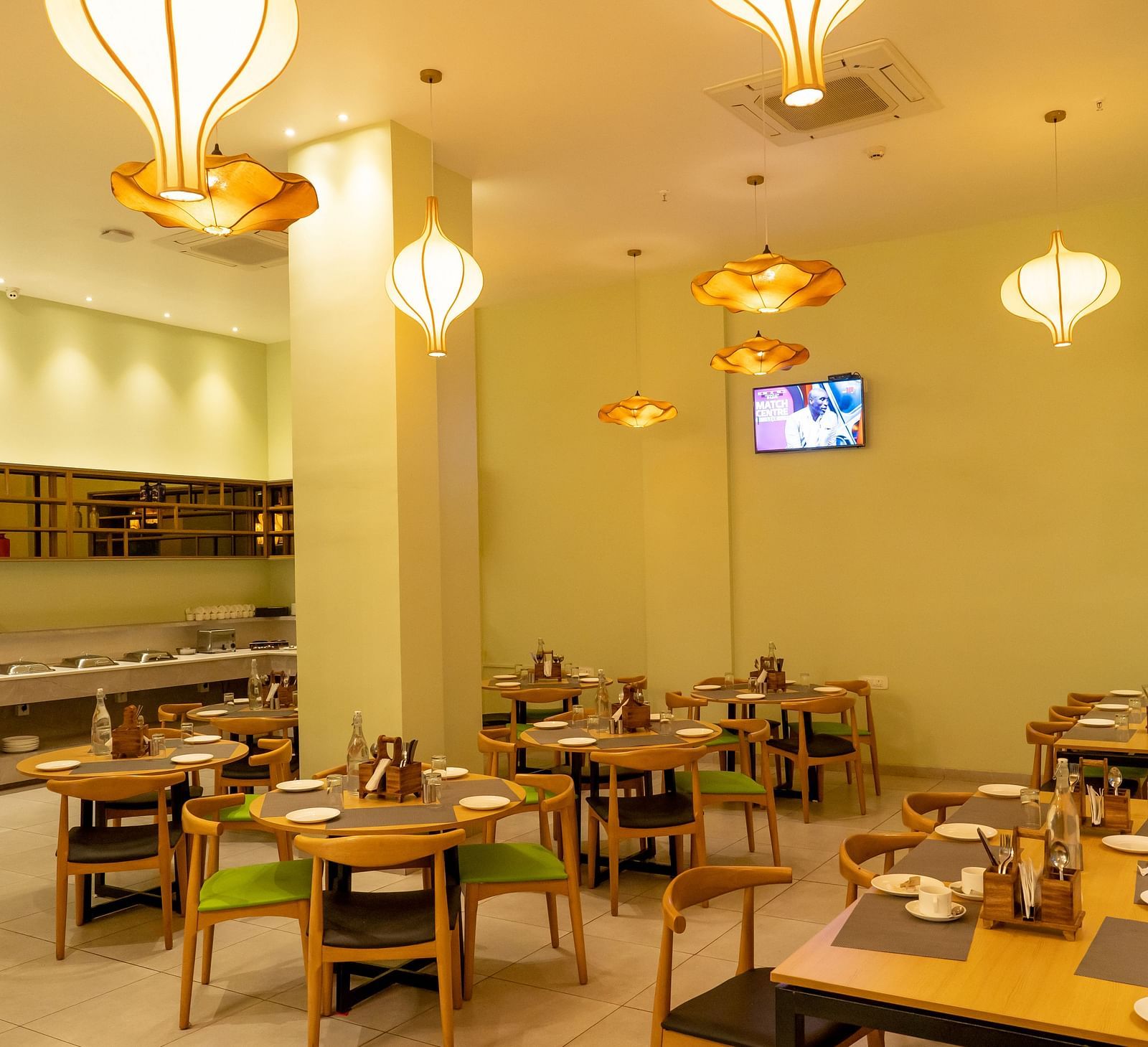 Another angle of the restaurant interior showing tables set for dining with hanging orange pendant lights and a TV screen on the wall - TGI Ameya Grand, Goa