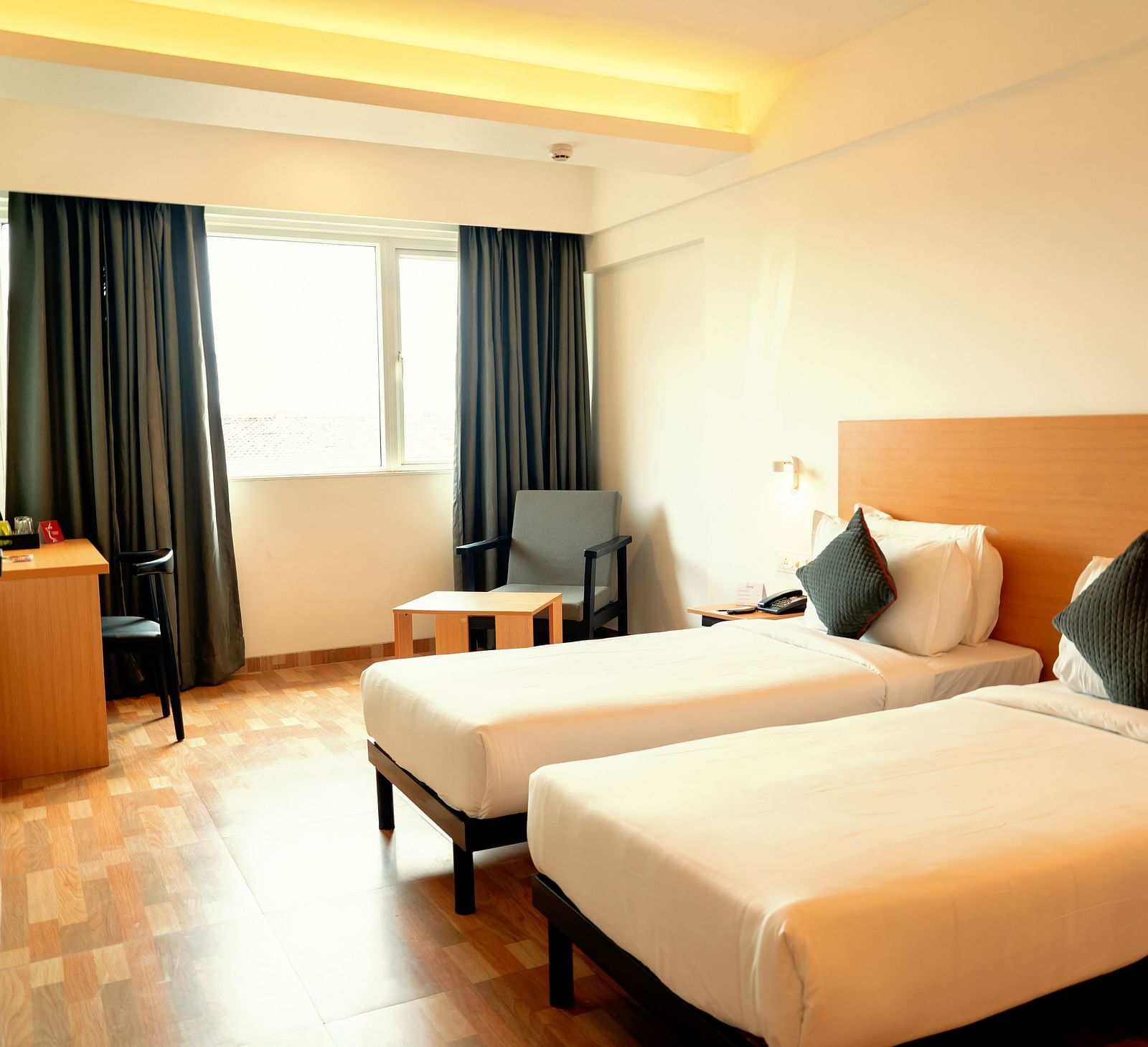 A bright hotel room with two single beds, a desk and a flat-screen TV, with a large window draped in gray curtains - TGI Ameya Grand, Goa