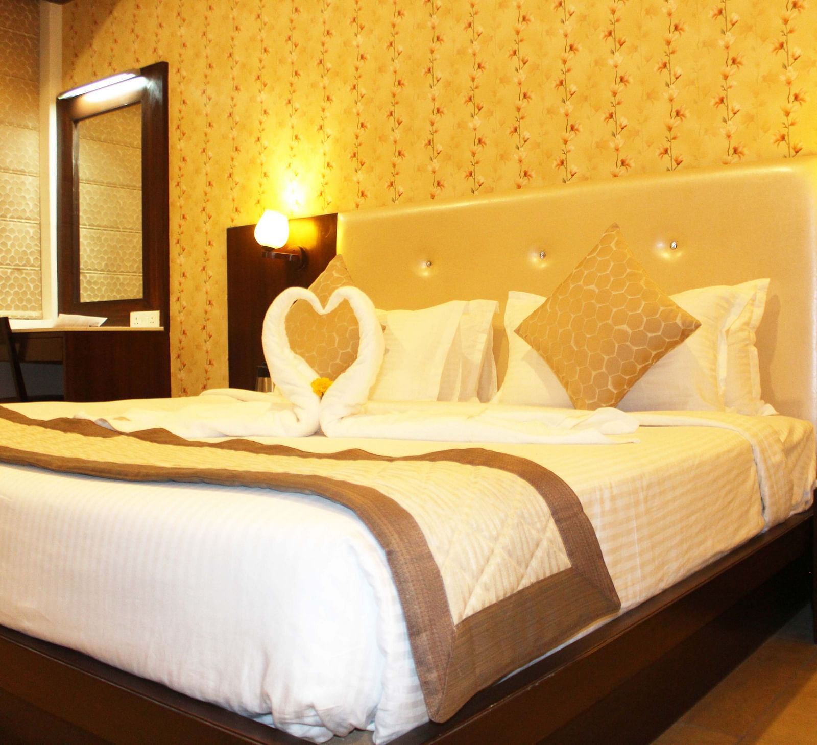 A warmly lit hotel room with a queen size bed, white bedding and a towel art swan on the bedspread - TGI Apple Inn, Jaipur 