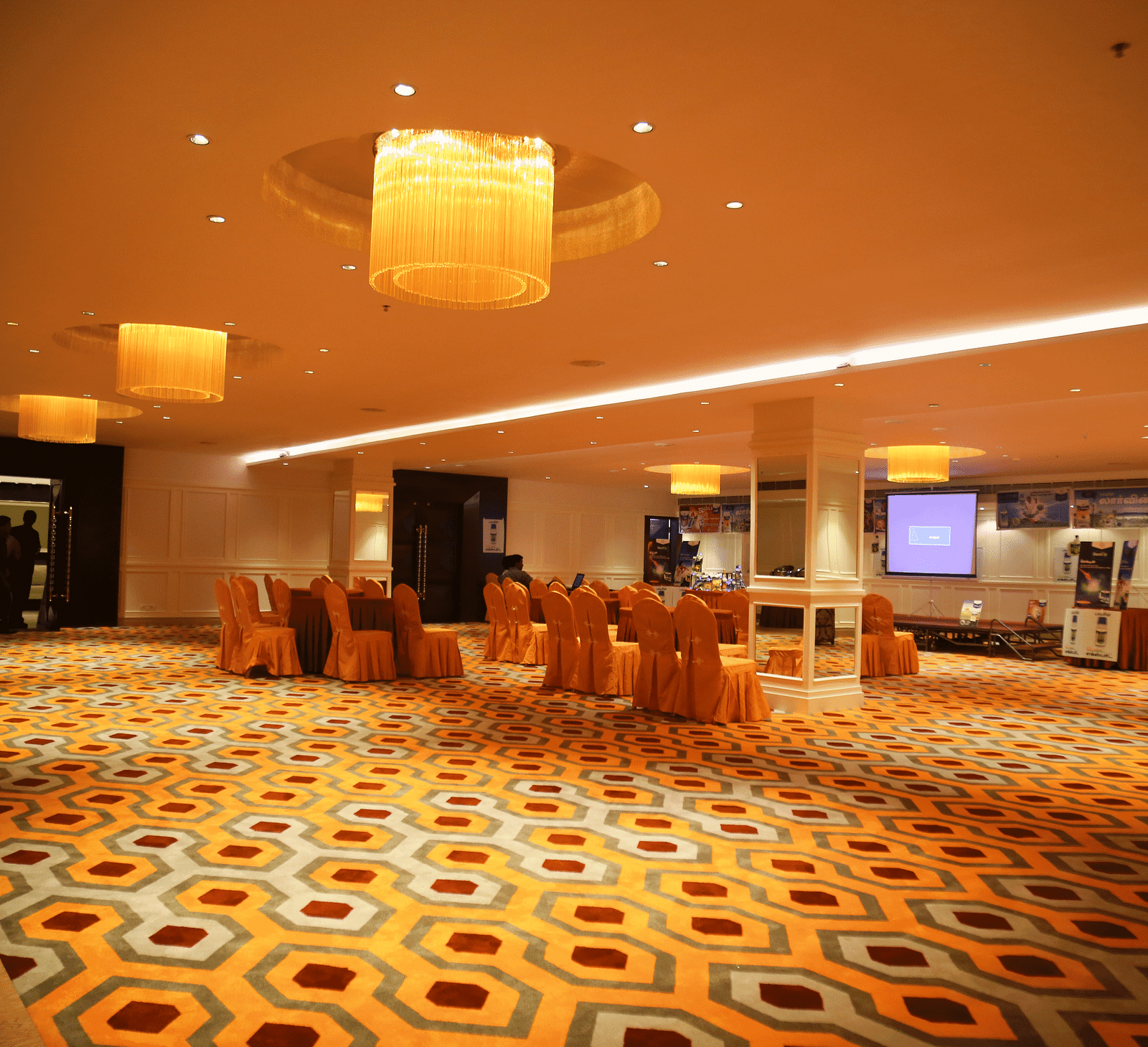 A banquet hall with orange chair covers and a central projection screen, under warm yellow lighting - TGI Grand Fortuna, Hosur