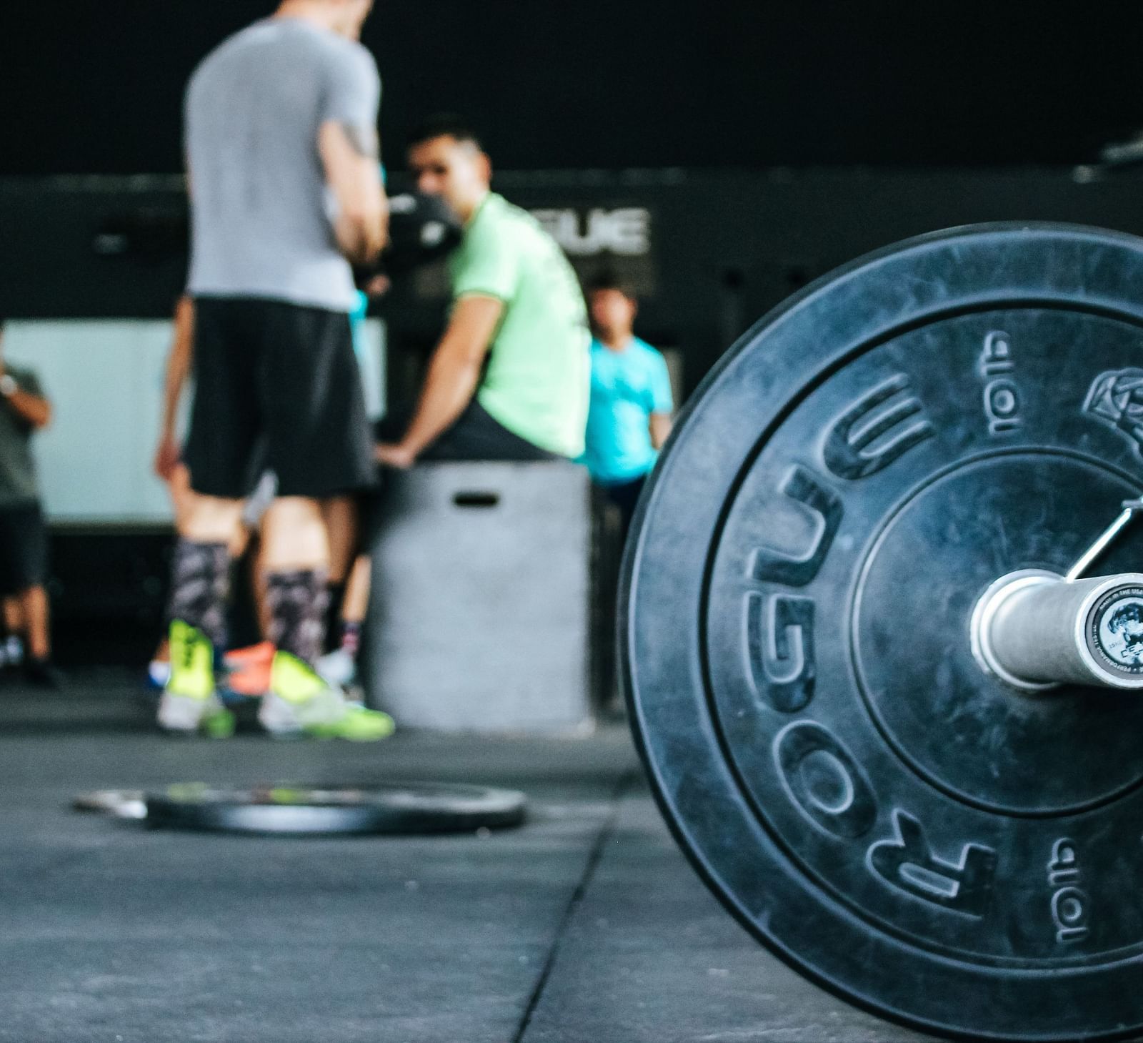 A gym scene focused on a barbell on the ground with out-of-focus people in the background