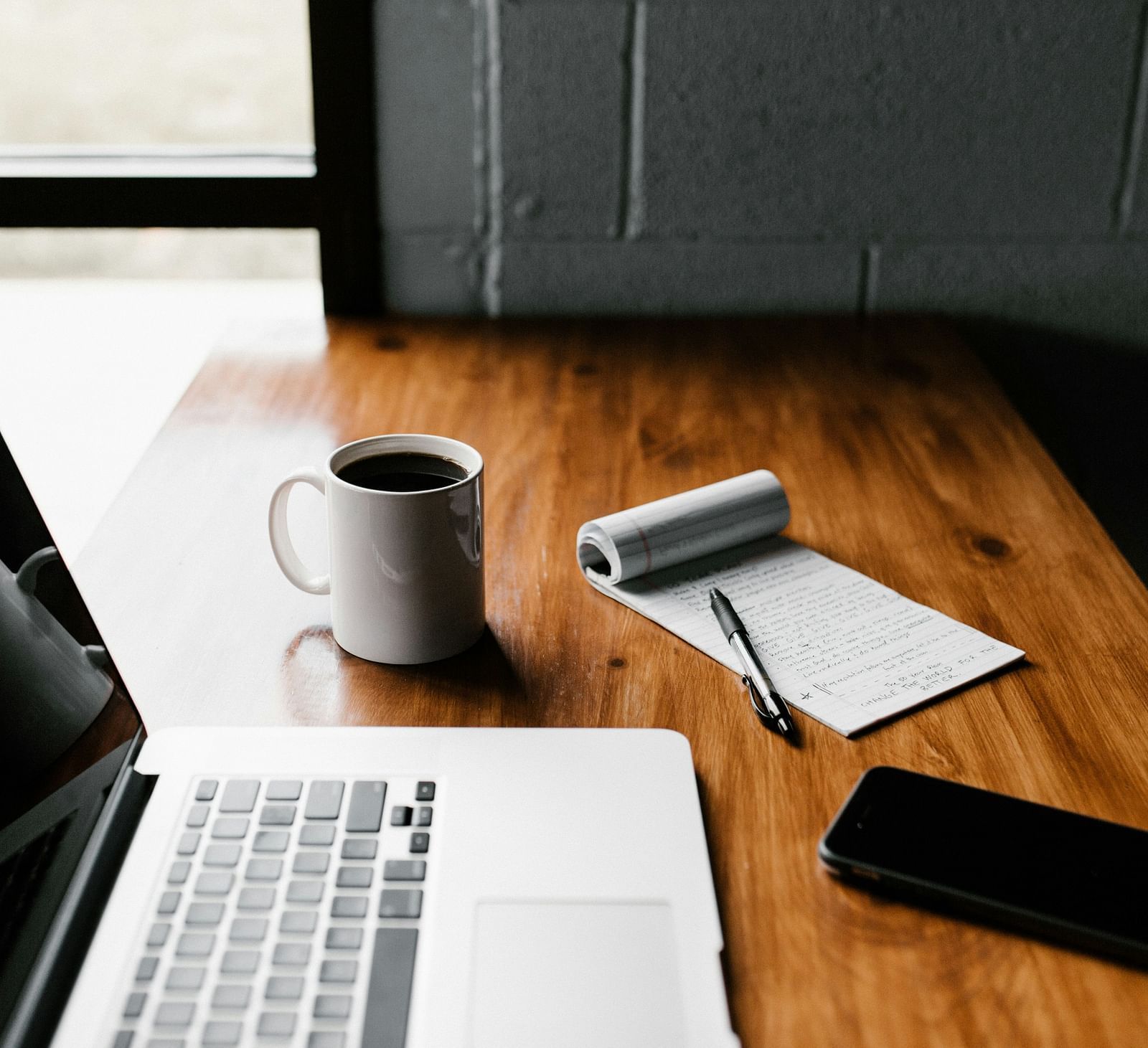 A laptop, smartphone, coffee cup, and notebook placed on a wooden desk