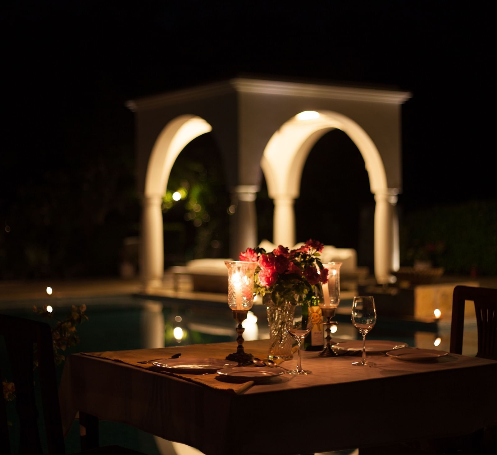 A dimly lit outdoor dining setup with a table set for two, featuring a flower arrangement and glassware, with a structure featuring arches in the blurred background- Shahpura Bagh.
