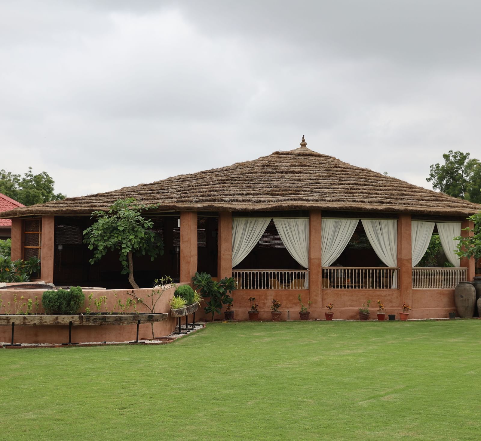The building at Surpura Bagh with a thatched roof, surrounded by a green lawn, lush plants, and a circular outdoor seating area.