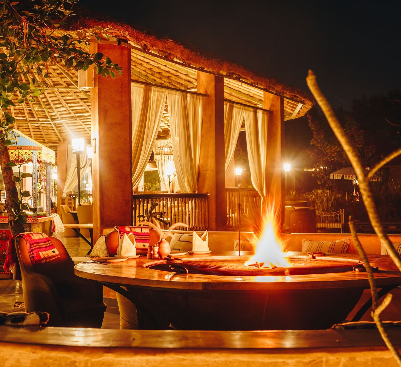 A view of a fire pit surrounded by chairs at Surpura Bagh, with warm lighting illuminating the thatched roof and decor.