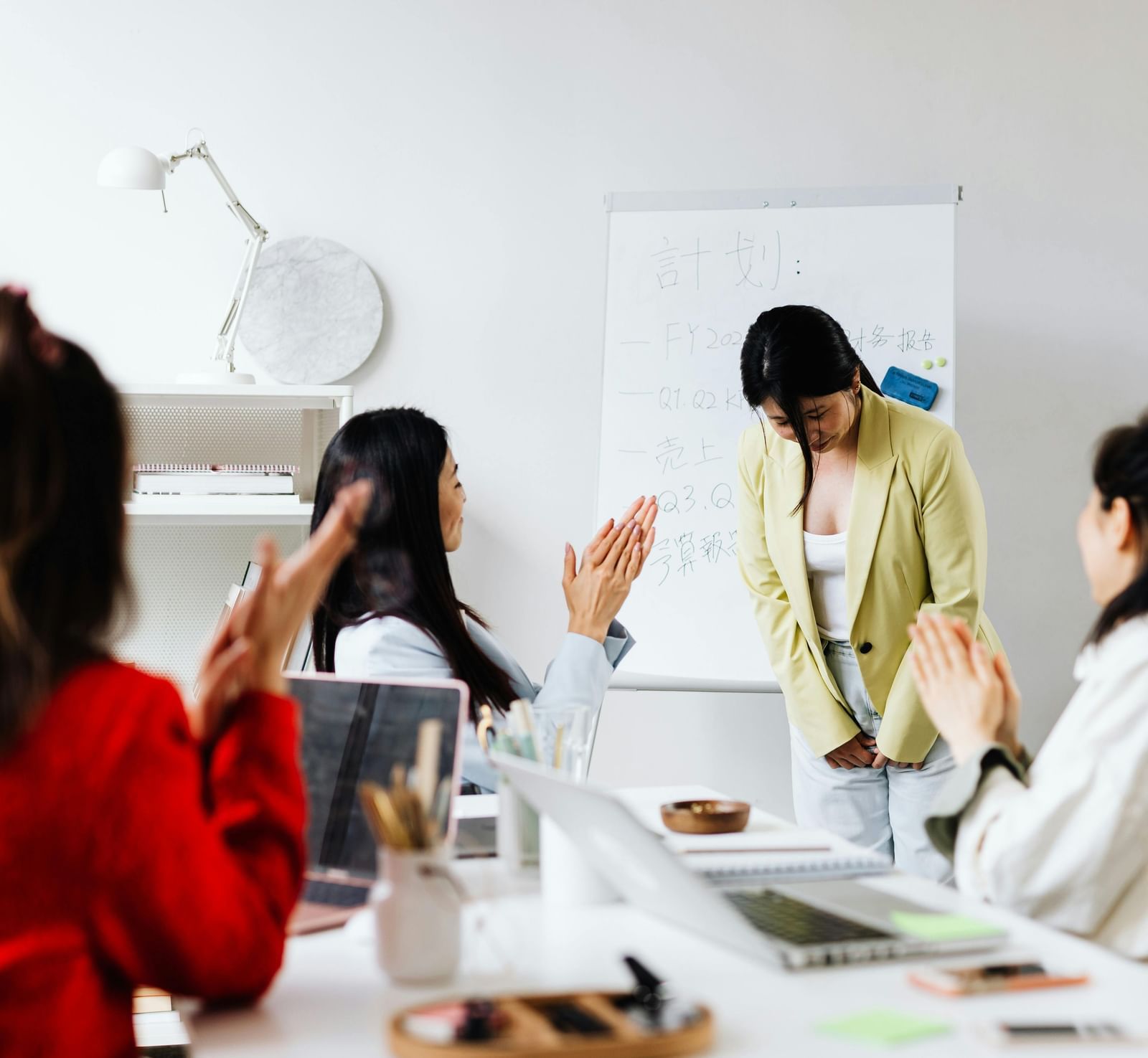 A brightly-lit workspace featuring people applauding to a team member as she bows down in gratitude.