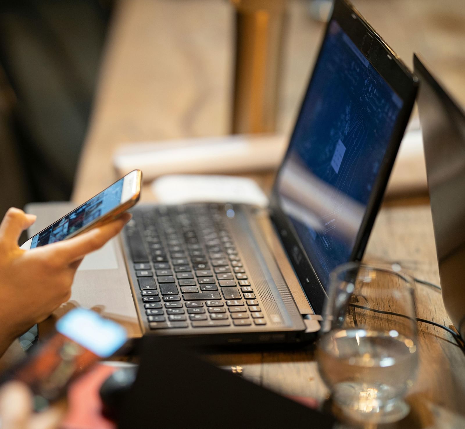 A laptop kept on a table beside a half-empty glass in front of a woman scrolling through her phone.