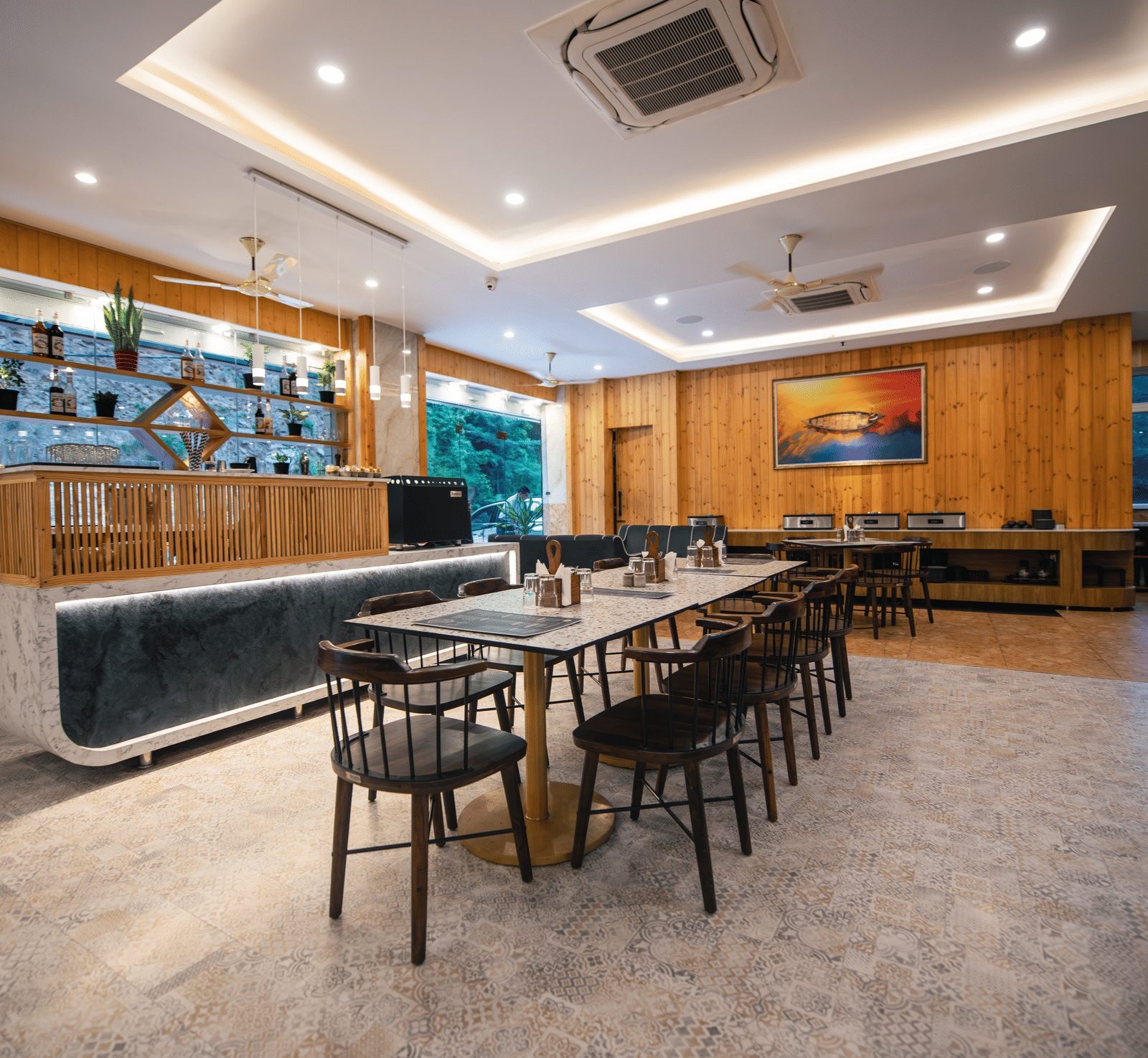 A restaurant at The Tattva Devaprayag with a long wooden table with black chairs and a bar.