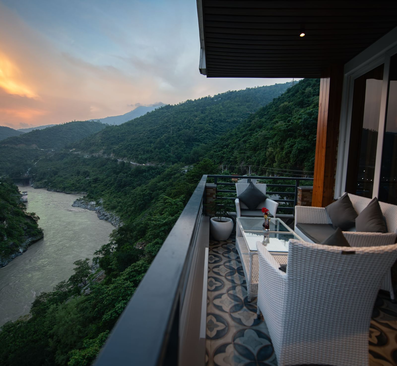  A wide view of the mountains and a river as seen from the balcony of The Tattva Devaprayag featuring a white couch and a table during sunset.