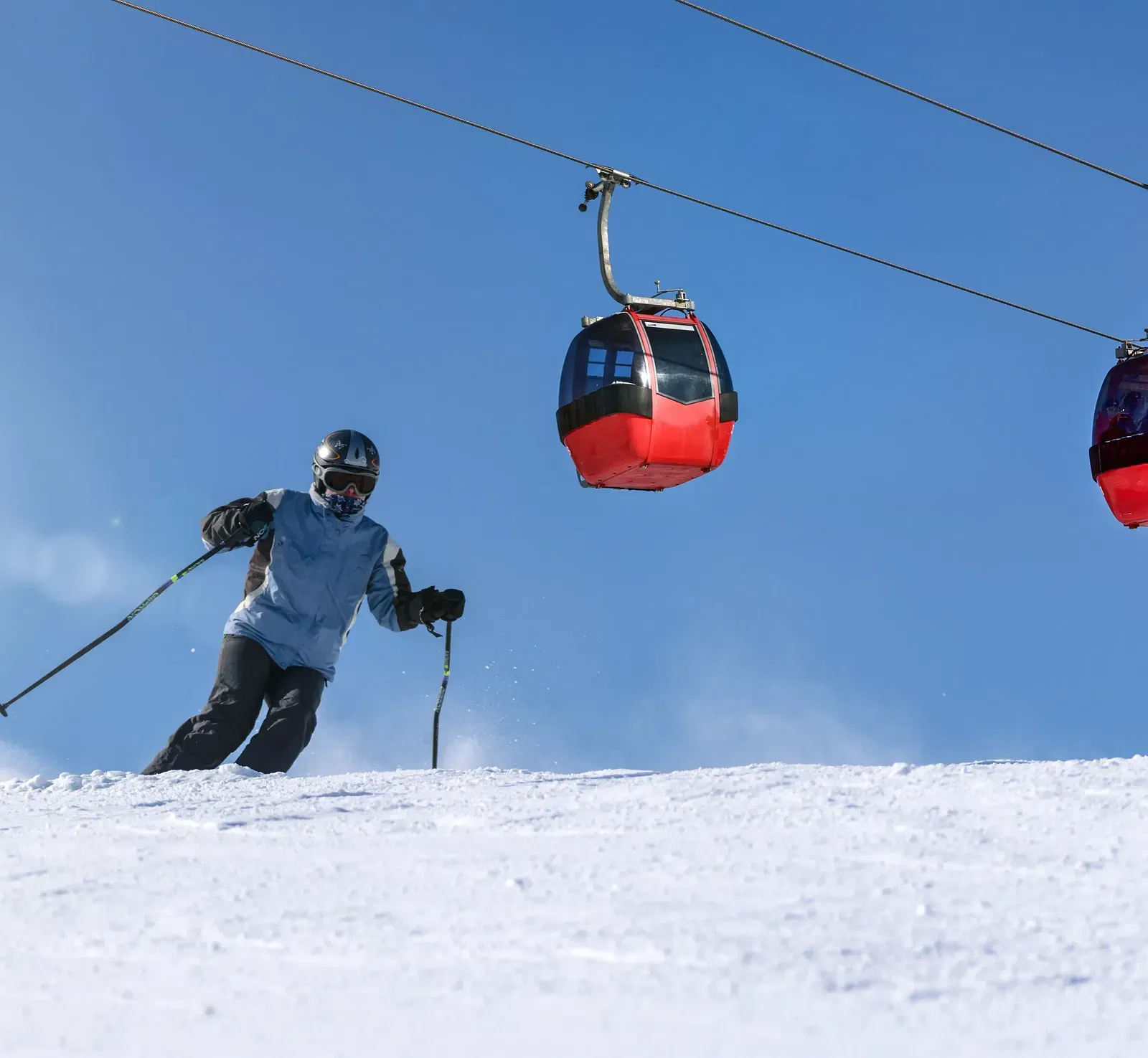 A skier descends a snowy slope with two red cable cars suspended above against a clear blue sky.