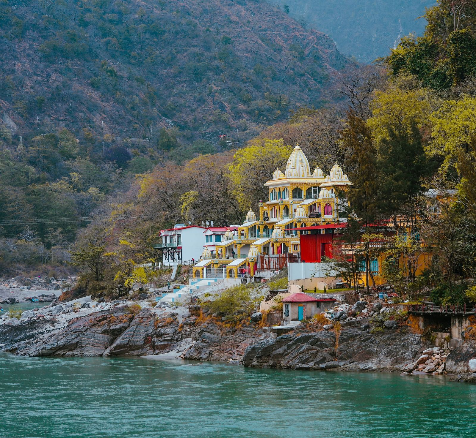 A brightly coloured temple complex resides by a tranquil river, tucked amongst verdant hills.