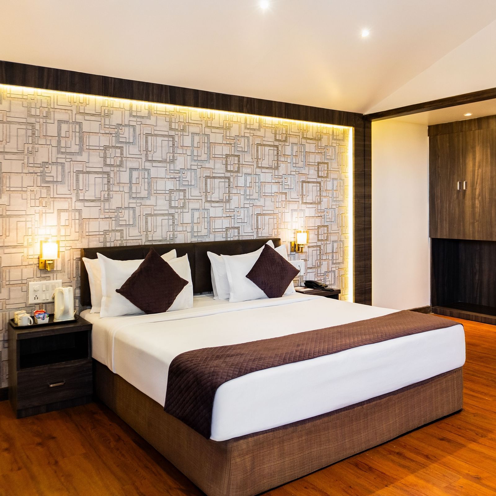 Modern hotel room with a king size bed, warm wooden flooring, geometric accent wall with soft backlighting, bedside tables, and cosy ambient lighting - The Grand Legacy Resort & Spa