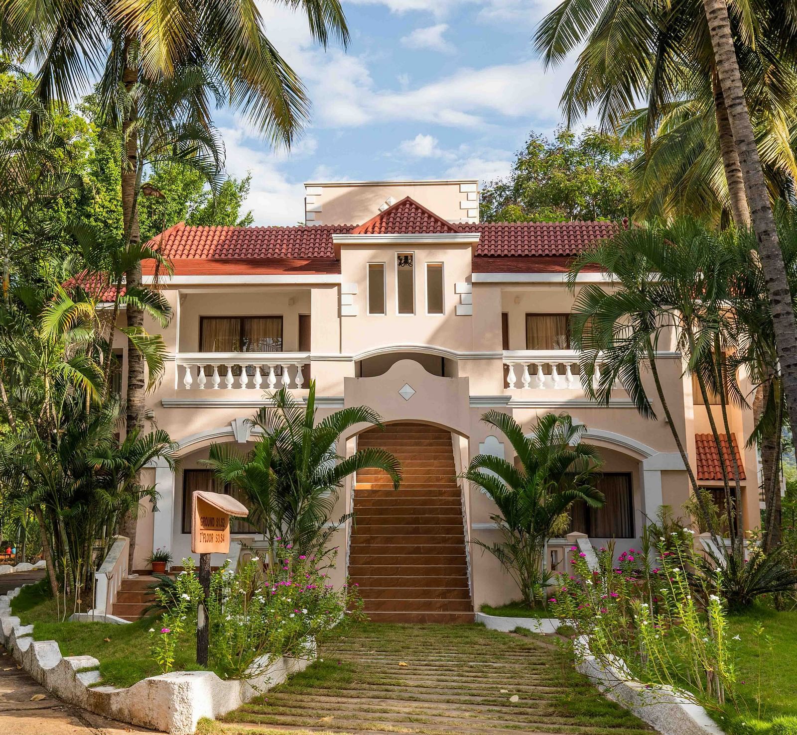 A grand two-storey villa-style building at Avana Resort with a terracotta roof, surrounded by palm trees and lush gardens under a bright sky.