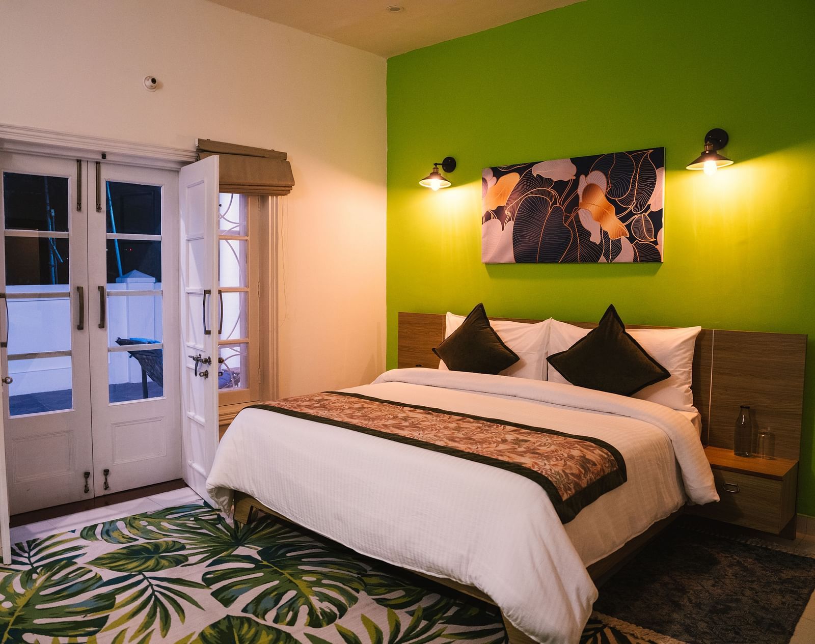 A spacious room at BluSalzz Villas - The Green Roof, Bhimtal, featuring a large bed with a photo frame above the headboard, night lamps glowing on either side, a carpet, and a door leading to the balcony.