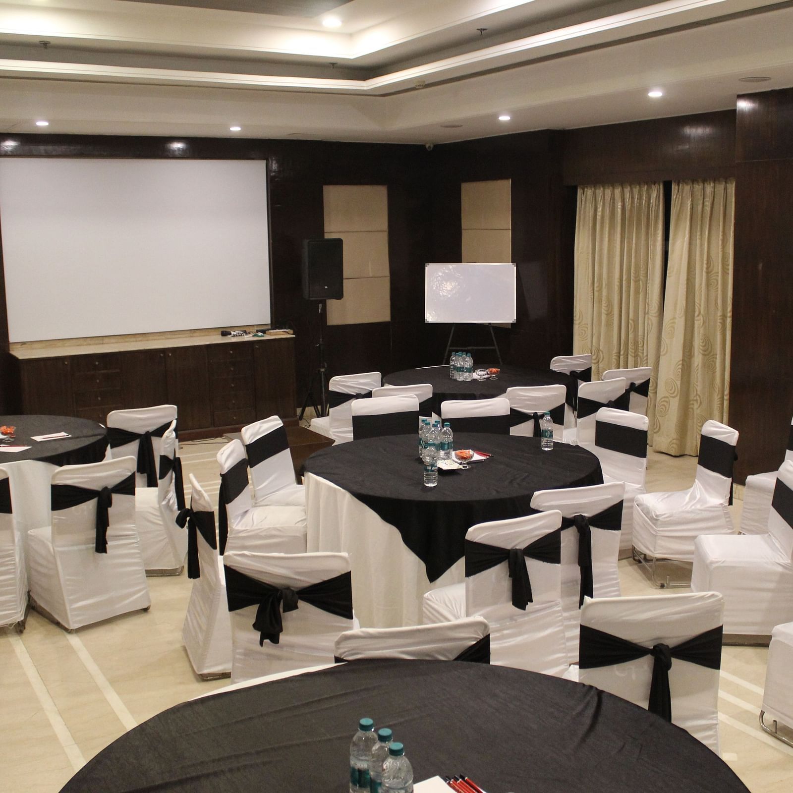 An image of a banquet hall at Inde Hotel Signature Tower, Gurgaon featuring a few round tables, each surrounded by chairs