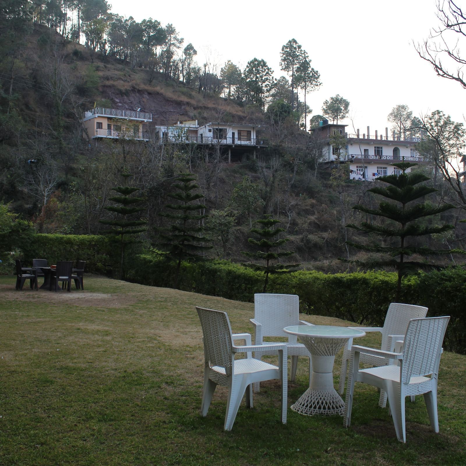 A beautifully maintained garden space with outdoor seating and a backdrop of hill slopes and trees at Dagshai Manor Kasauli - Inde Hotels.
