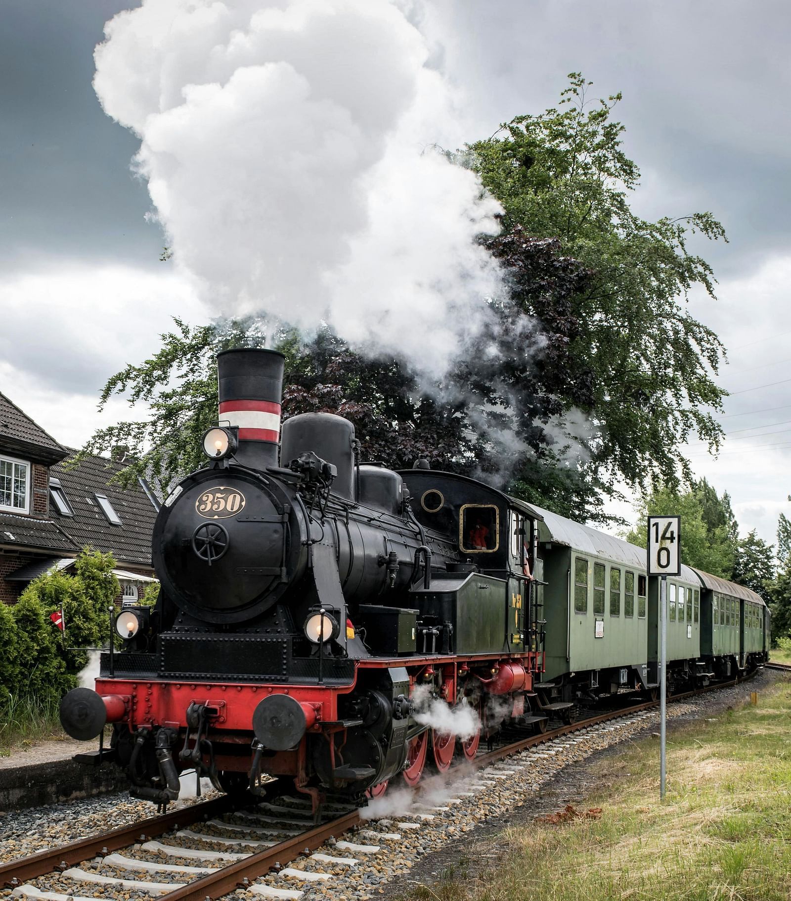 A vintage steam train passing by houses under a cloudy sky.