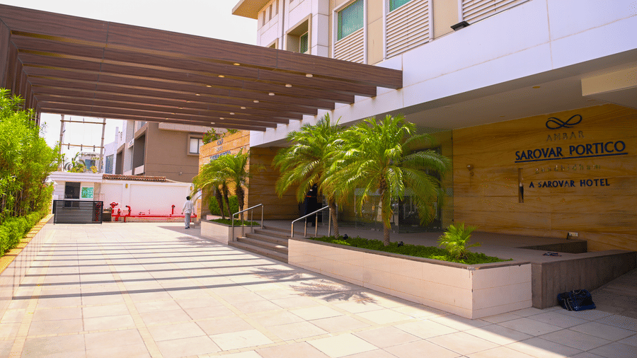 A daylight view of the main building exterior at Ambar Sarovar Portico, Gandhidham showing a covered canopy entrance, steps, and small palm trees.