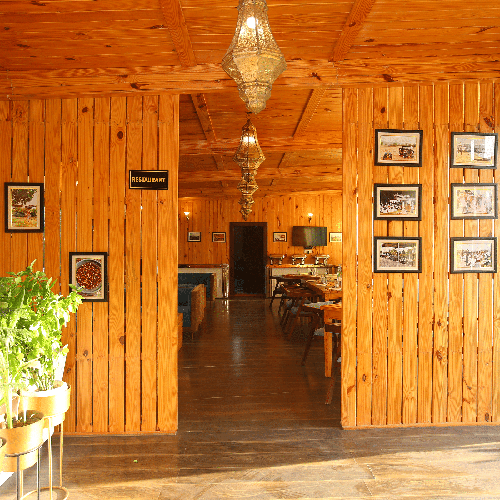 Entryway to the dining area featuring wall-mounted photo frames, a hanging lamp, and potted plants on the side at Aanandam Resort & Spa