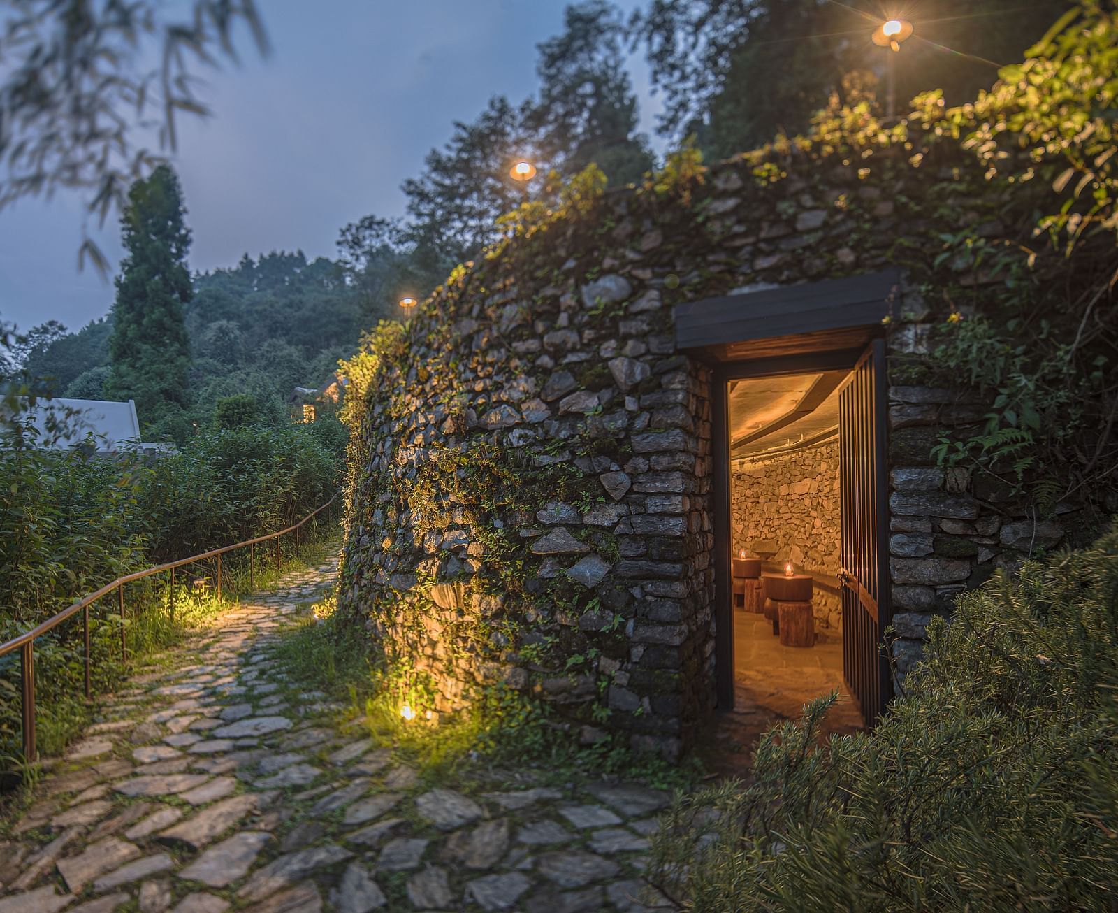 Stone pathway leading to a warmly lit room entrance, surrounded by lush greenery.