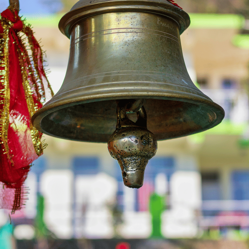 Close-up of a traditional brass temple bell decorated with red prayer threads hanging outdoors with a softly blurred background.