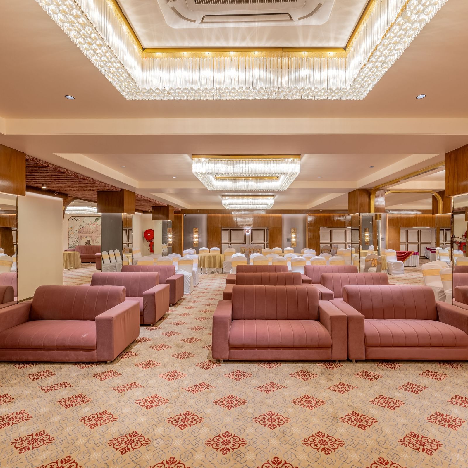 A spacious banquet hall with maroon sofas, round tables and decorative lighting on the ceiling at Hotel Highway King, Jaipur.
