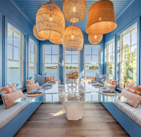 A vibrant, sunlit dining room featuring floor-to-ceiling windows and blue walls. A glass-topped table is surrounded by built-in bench seating with gray cushions and colorful patterned pillows.