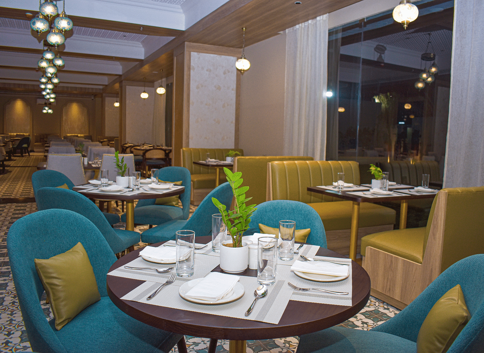 A restaurant interior with teal armchairs, booth seating, round tables, a patterned floor, sheer curtains, and hanging pendant lights - Urban Suites by BluSalzz, Jaipur