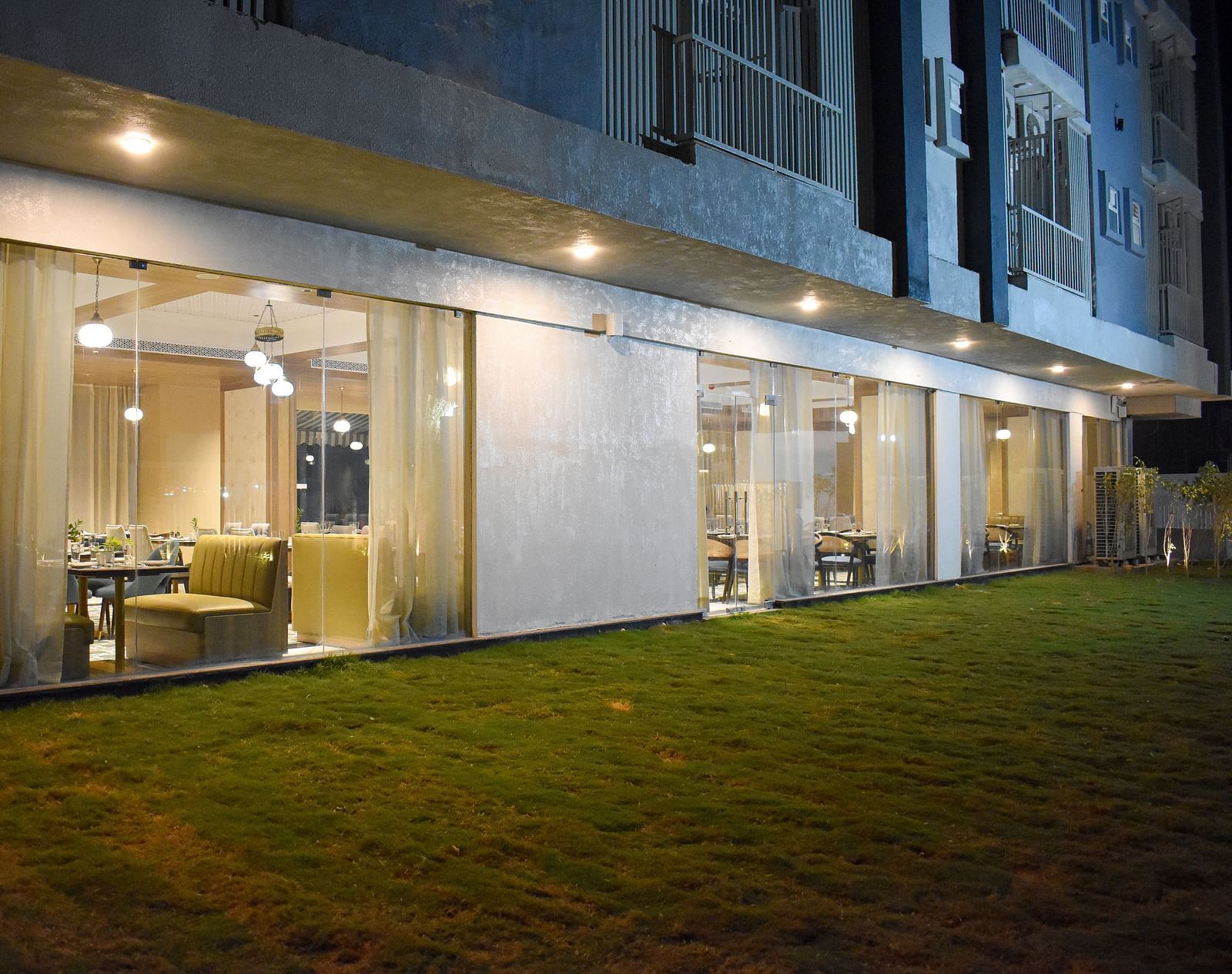 Exterior view of a building with a glass-walled restaurant, sheer curtains, hanging lights, and a grassy area in front - Urban Suites by BluSalzz, Jaipur