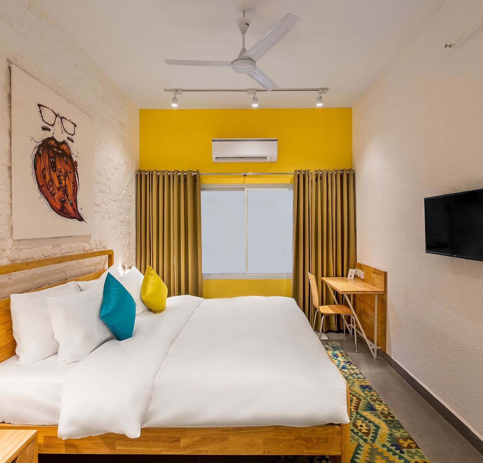 An side angle interior image of the standard room at Bedzzz Varanasi showcases a spacious layout, featuring a comfortable double bed, a cozy floor mat, a stylish study table and chair, a charming coffee table, elegant curtains, an air conditioner for a ref