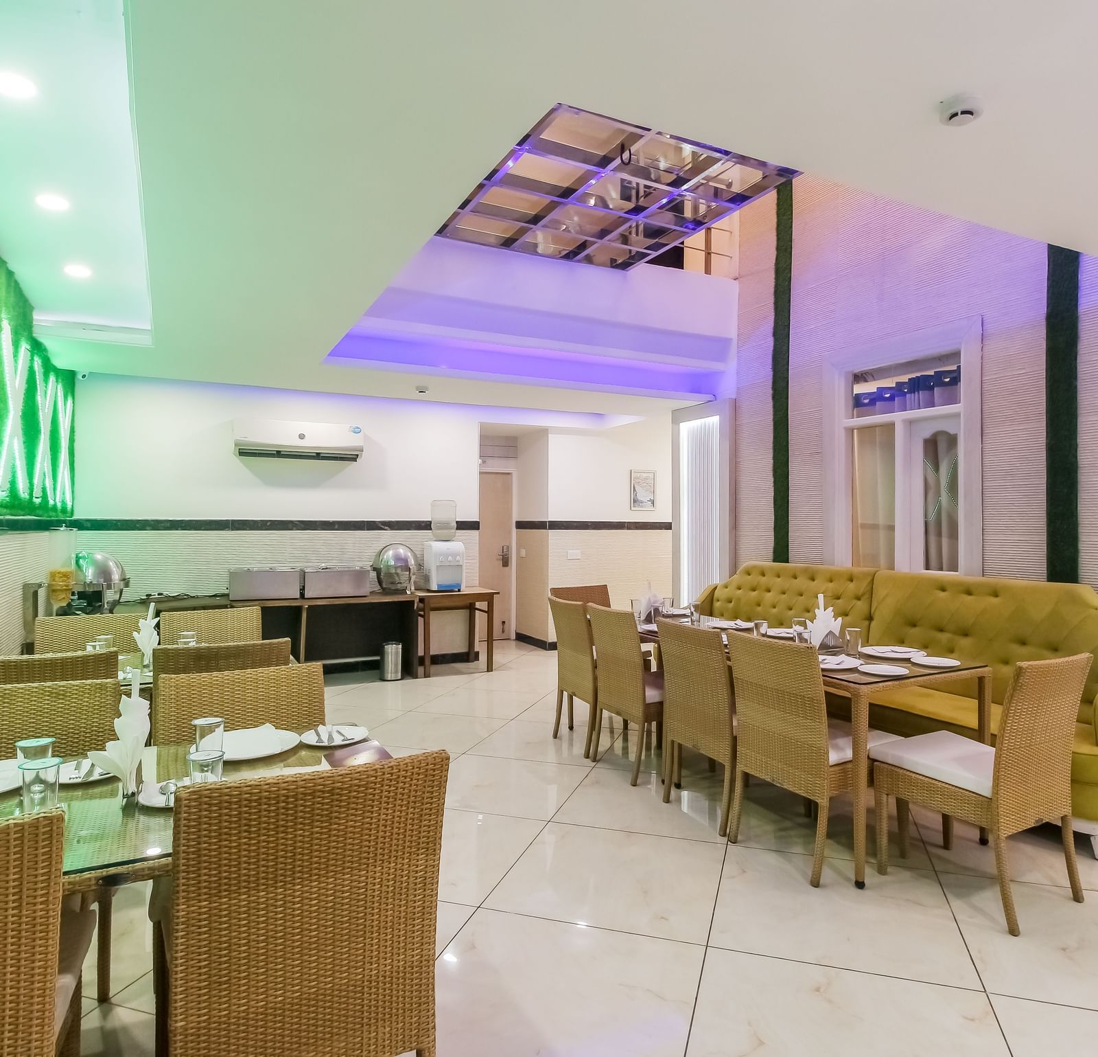 A restaurant interior with a long tufted sofa, wooden tables and chairs, a decorative wall design, and a tiled floor, lit by soft lighting. - Hotel Diamond Plaza, Chandigarh.