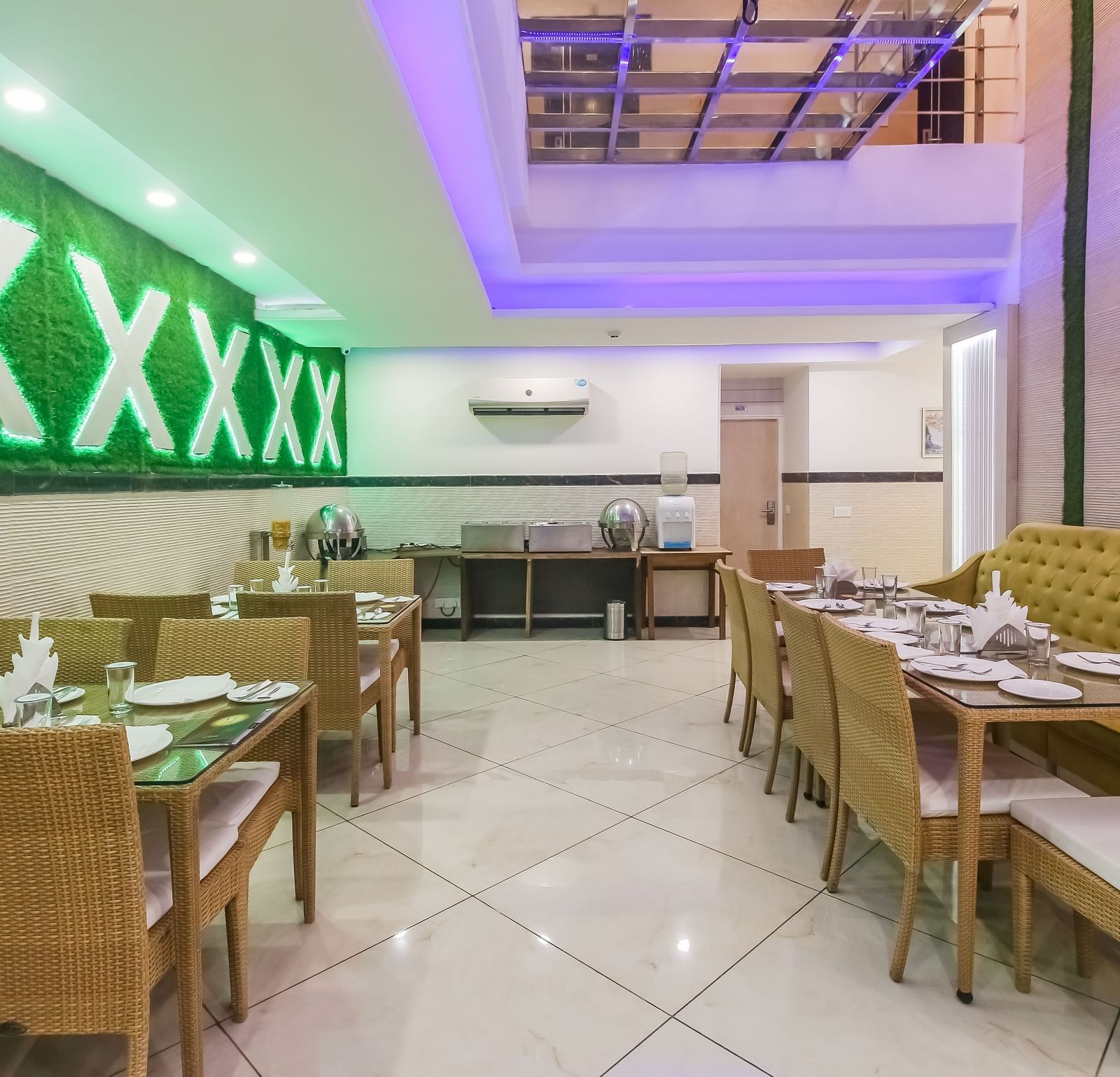 A restaurant interior with a long tufted sofa, wooden tables and chairs, a decorative wall design, and a tiled floor, lit by soft lighting. - Hotel Diamond Plaza, Chandigarh.