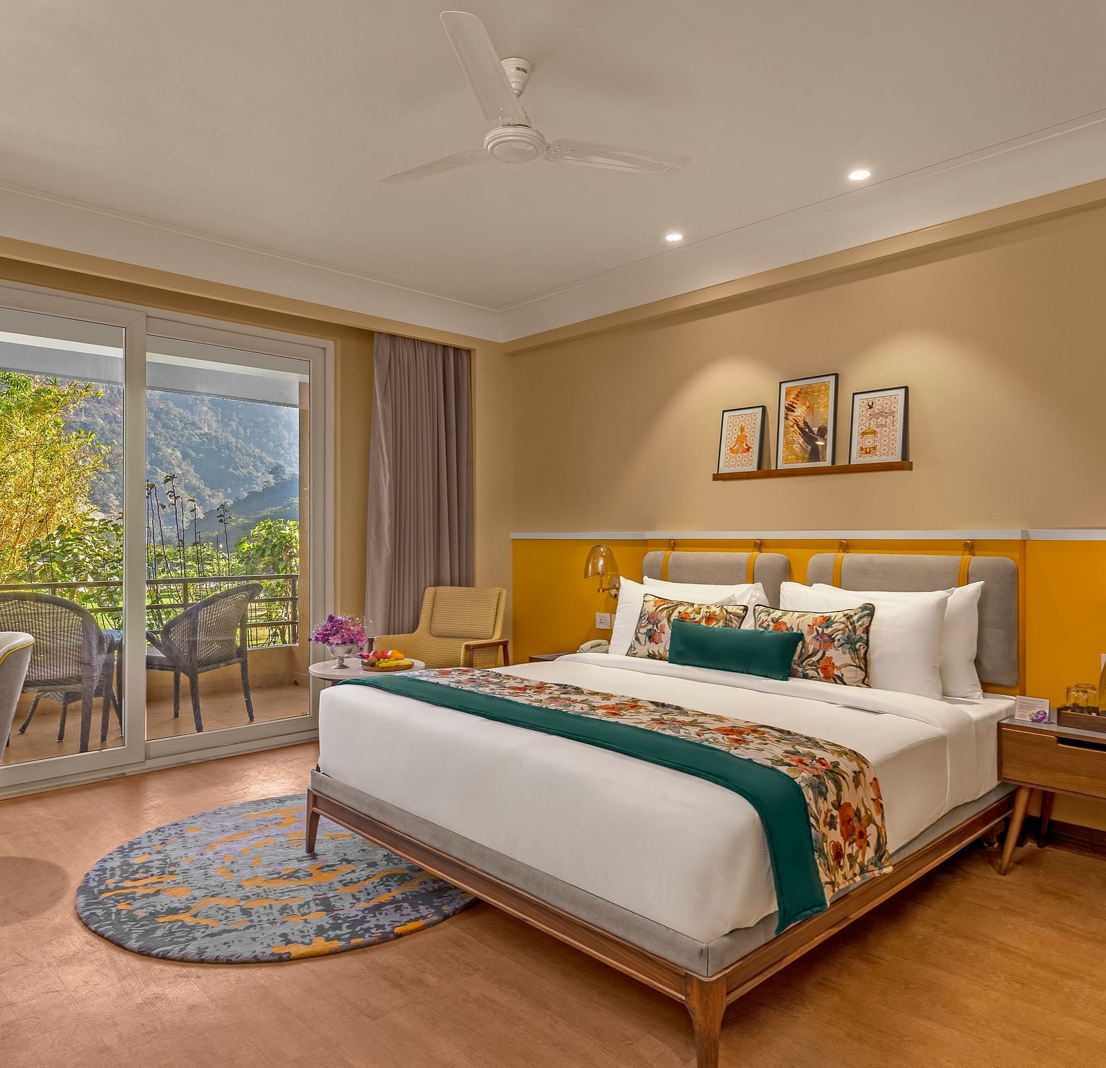 Image of a  room features a large king-size bed with a plush headboard and comfortable bedding. A comfortable armchair sits by a large window offering breathtaking views. The room also includes a wooden desk with a chair, a television, and a ceiling fan. T