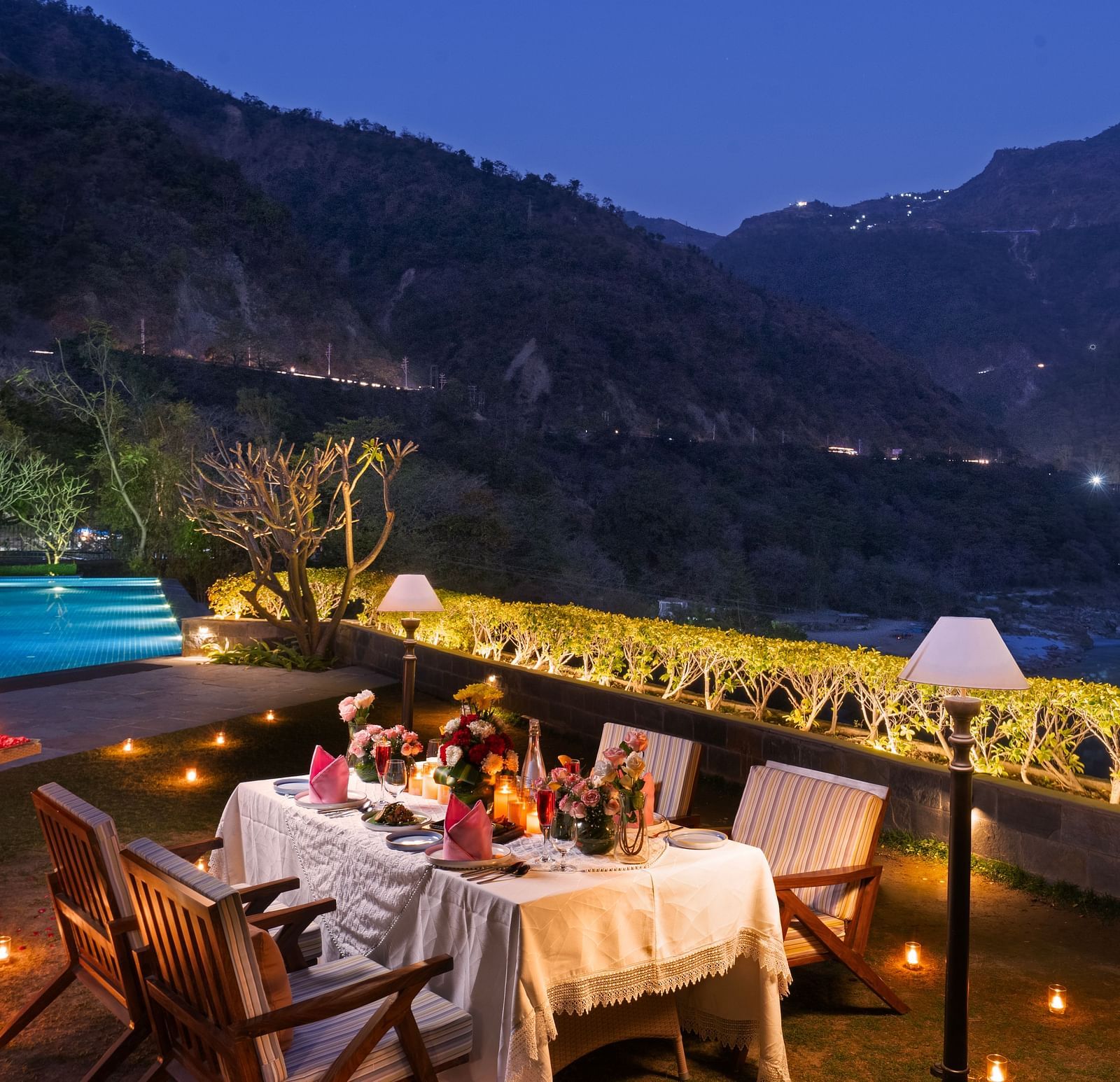 A dinner setup by a pool with mountains in the background. The table is adorned with candles and flowers.