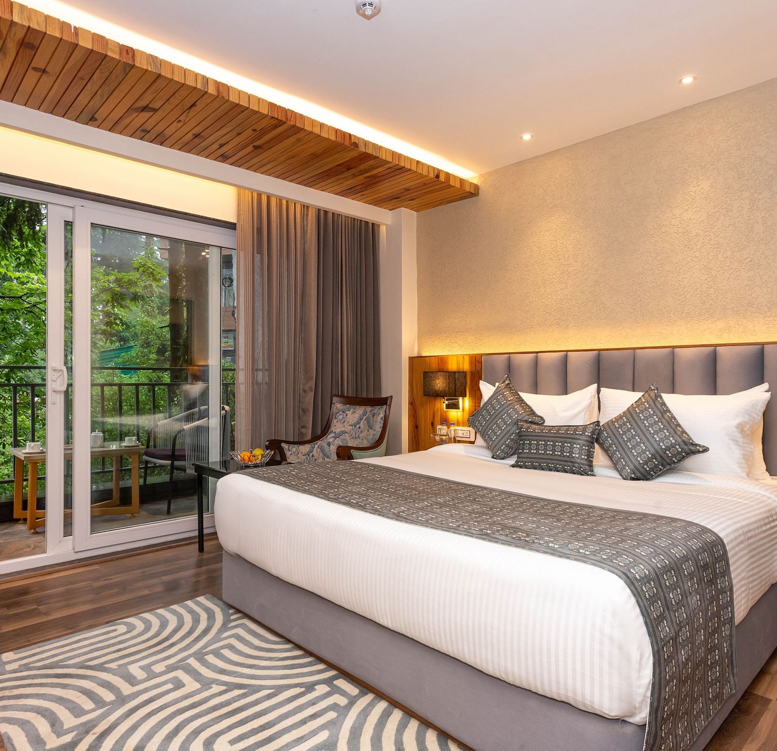 An image of a Superior Room with bed, bed side table, lamps, rug, and an expansive window leading to the balcony - The Belvedere Himalayan Retreat, McLeodganj