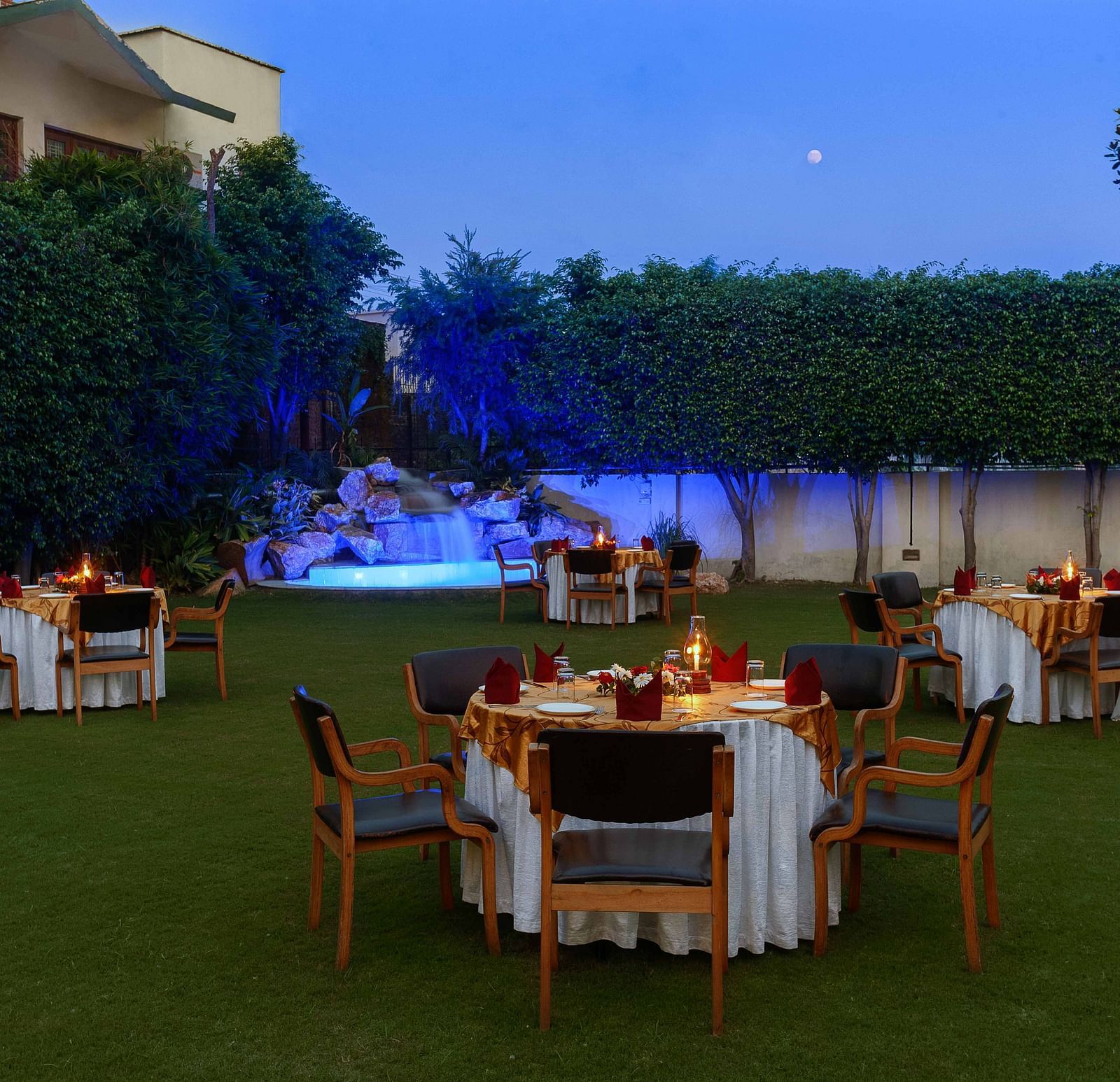 An open lawn with tables and chairs arranged in a circle, each adorned with decorative settings and a small lamp, featuring a lit-up fountain in the corner