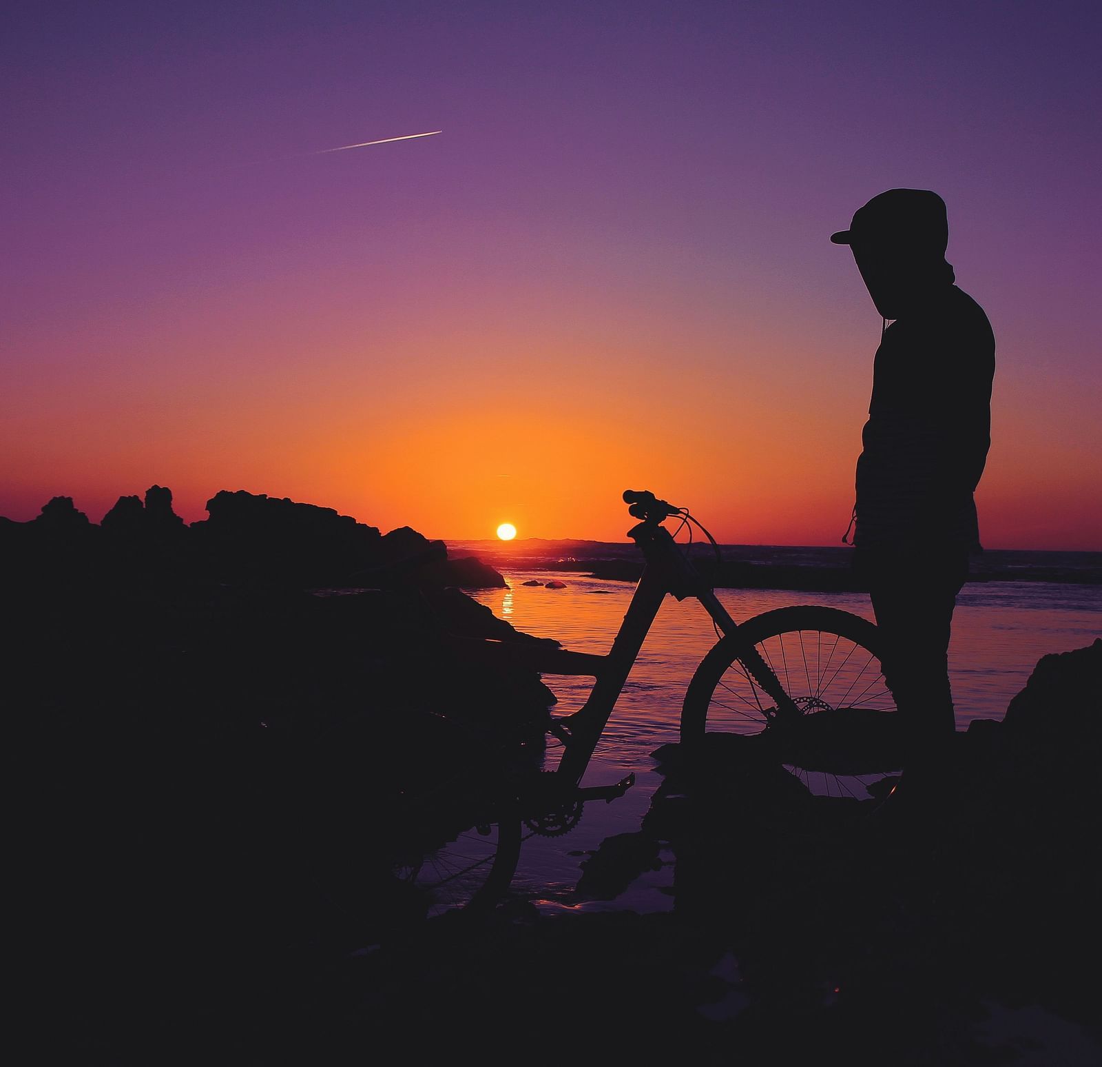 A person looking at the beach and the sunset while standing next to a bicycle.