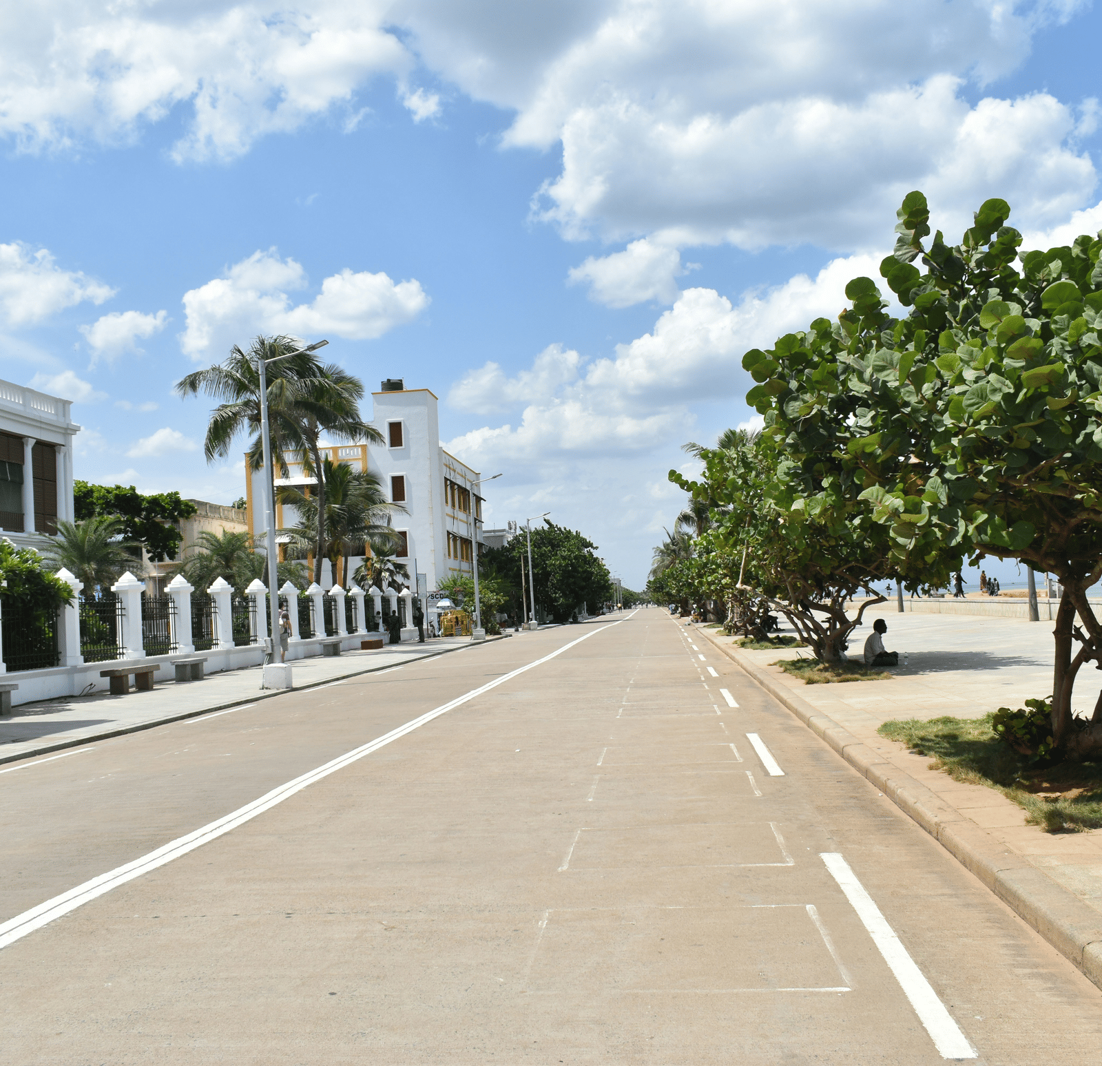 A wide, empty road lined with a white building and trees under a bright blue sky.