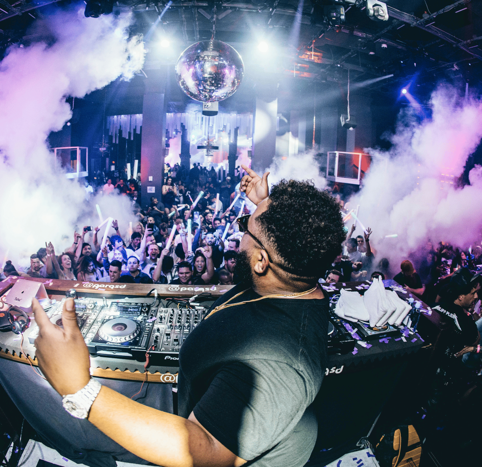A view from behind the stage of a DJ performing for a crowd with strobe lights on it.