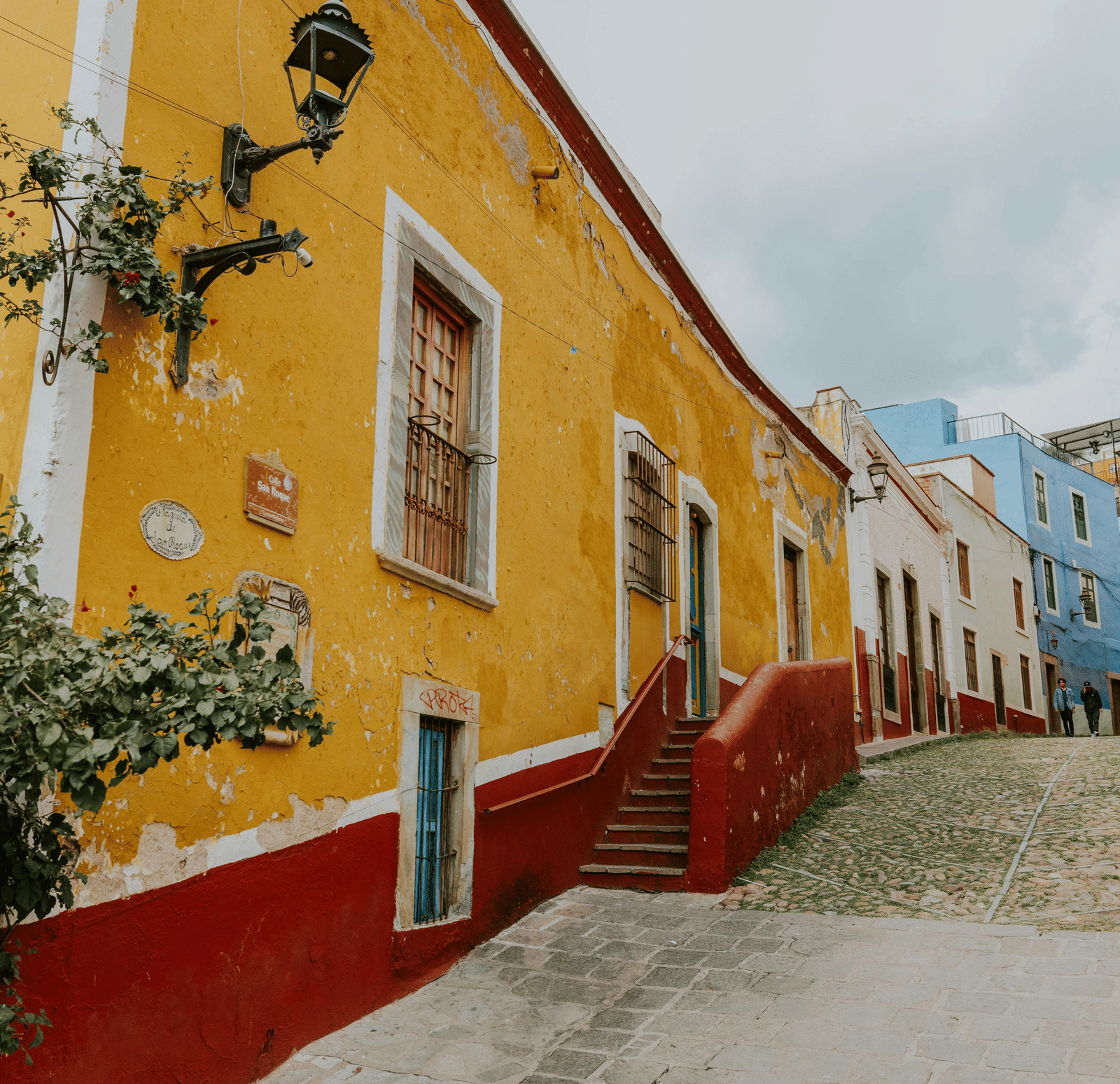 A street corner with a red and yellow building on the left and blue and white buildings in the distance is shown from a low angle | French Colony Pondy