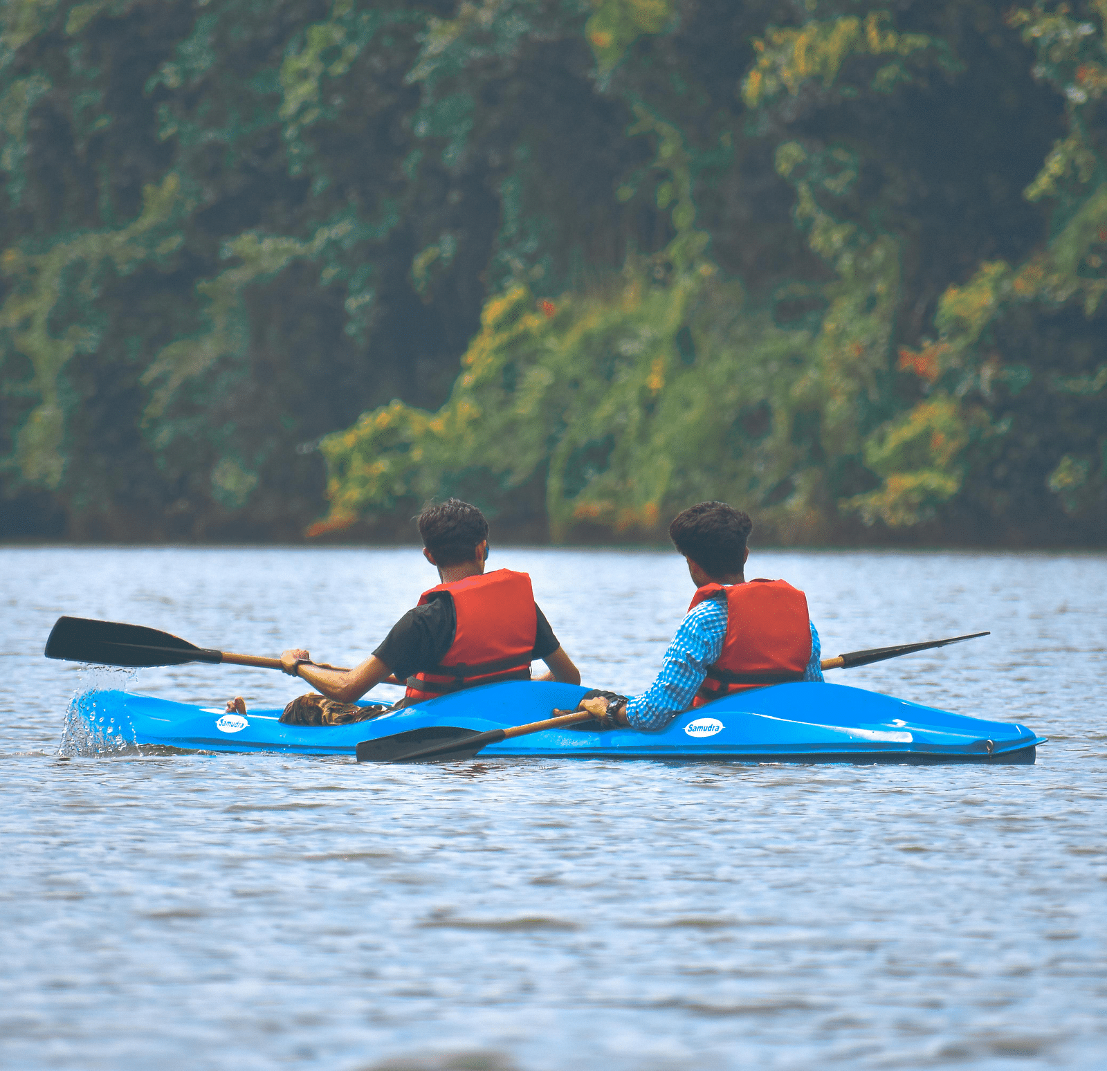 Two people wearing life jackets paddle a blue kayak on a calm lake surrounded by trees.